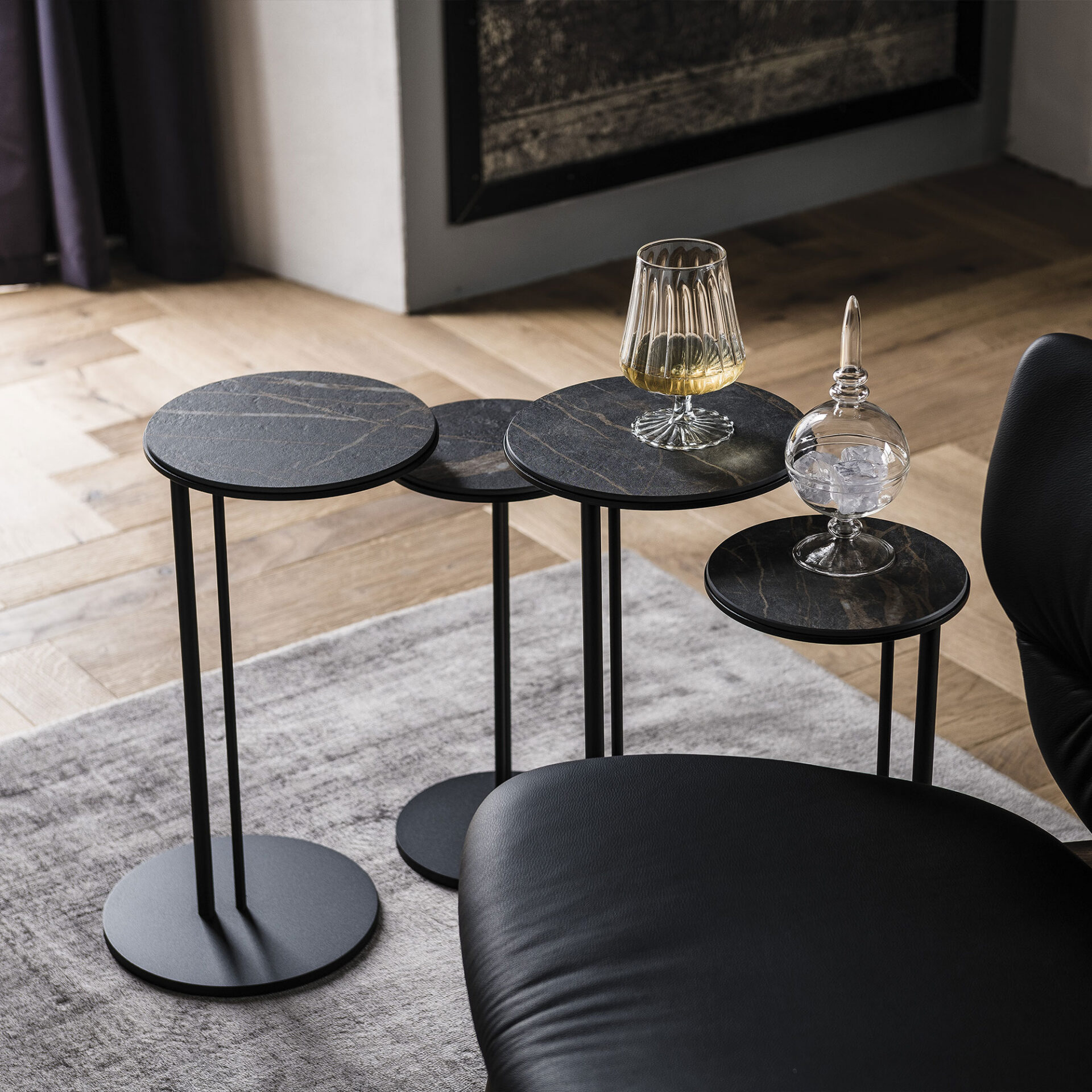 Four modern nesting side tables with dark marble tops and black metal bases, arranged on a gray rug. One table holds a glass with a golden drink, another a decorative glass filled with ice. A black leather armchair is partially visible.