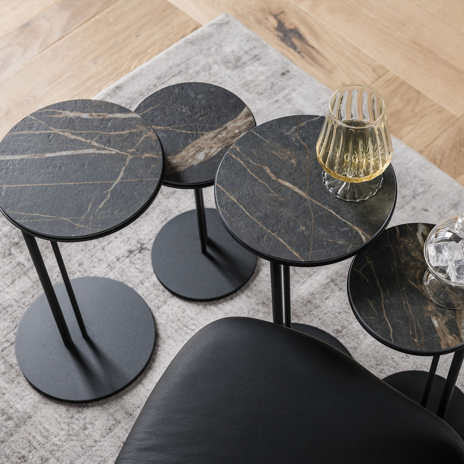 Overhead view of four modern, round side tables with dark, marble-patterned tops and black metal bases, arranged on a grey rug and wood floor. One table holds a ribbed glass with a drink.