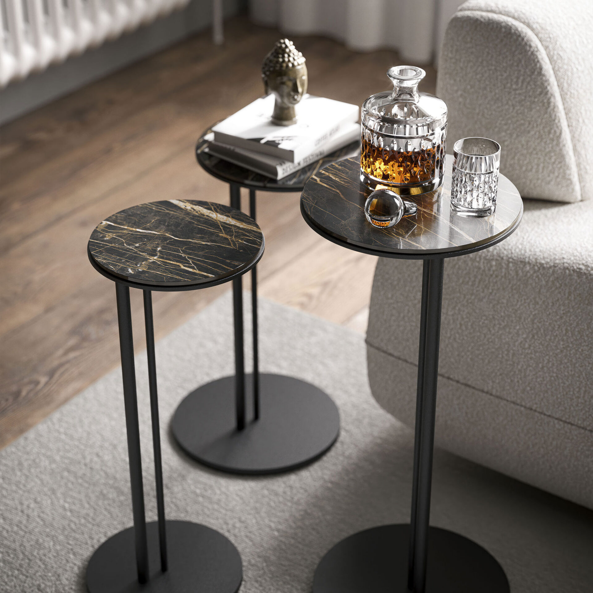 Three modern side tables with black and gold marble tops and slender black legs. On them are a crystal decanter with amber liquid, a glass, books, and a Buddha statue. A light grey rug and sofa are in the background.