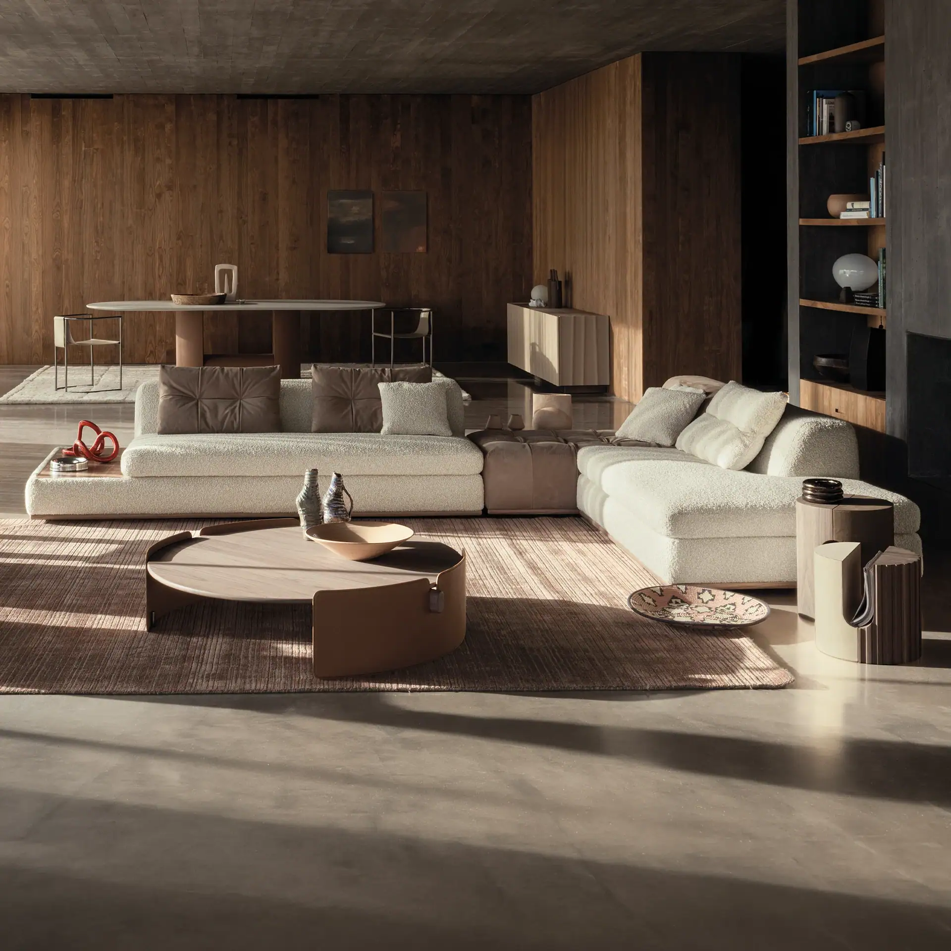 A modern living room with an L-shaped off-white bouclé sofa, round wooden coffee table, and a dining area with an oval table, all set against wooden panel walls and a concrete floor with sunlight streaming in.