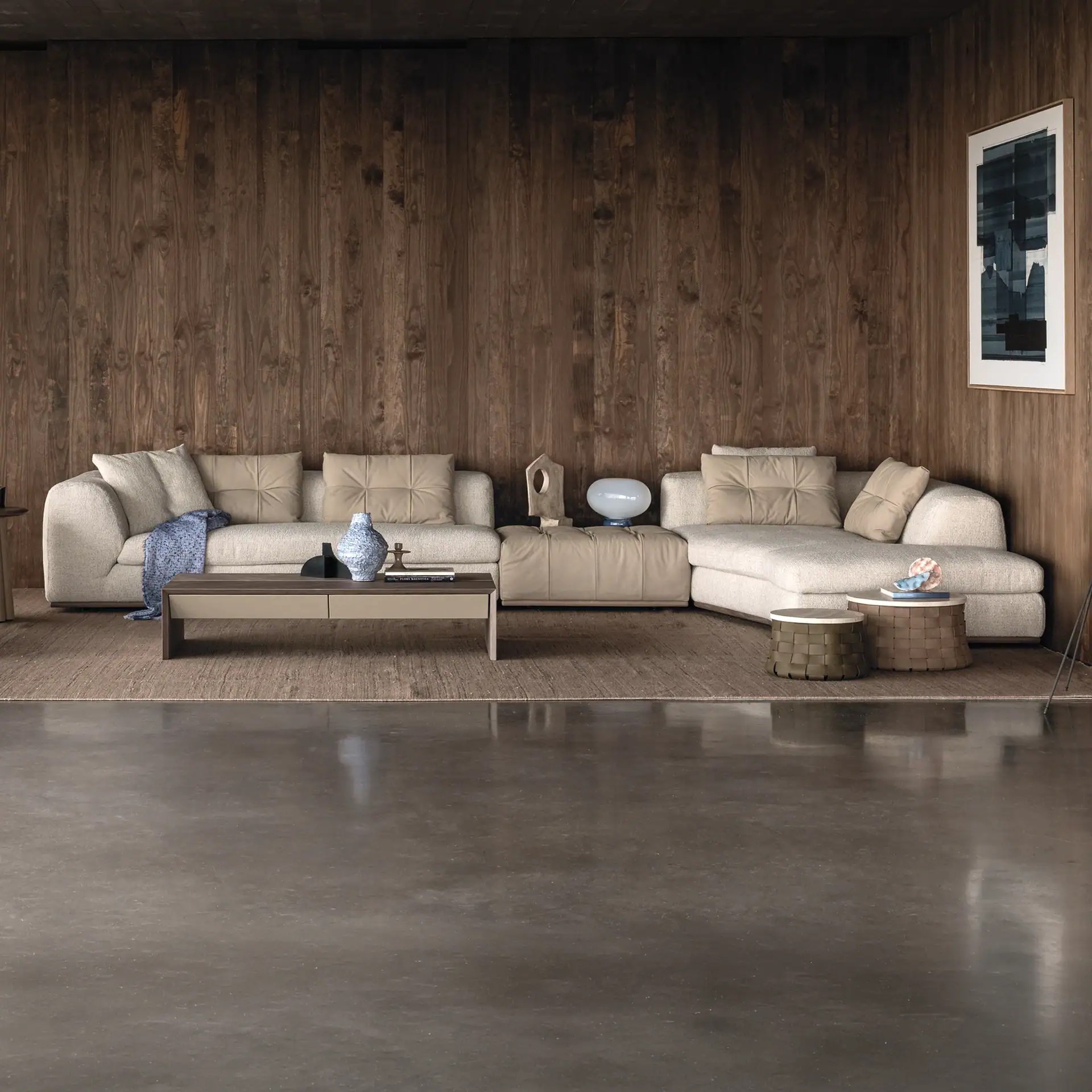 A modern living room with an L-shaped beige sofa, dark wood paneled walls, and a polished concrete floor. A coffee table and two woven side tables are adorned with decorative items, and an abstract artwork hangs on the wall.