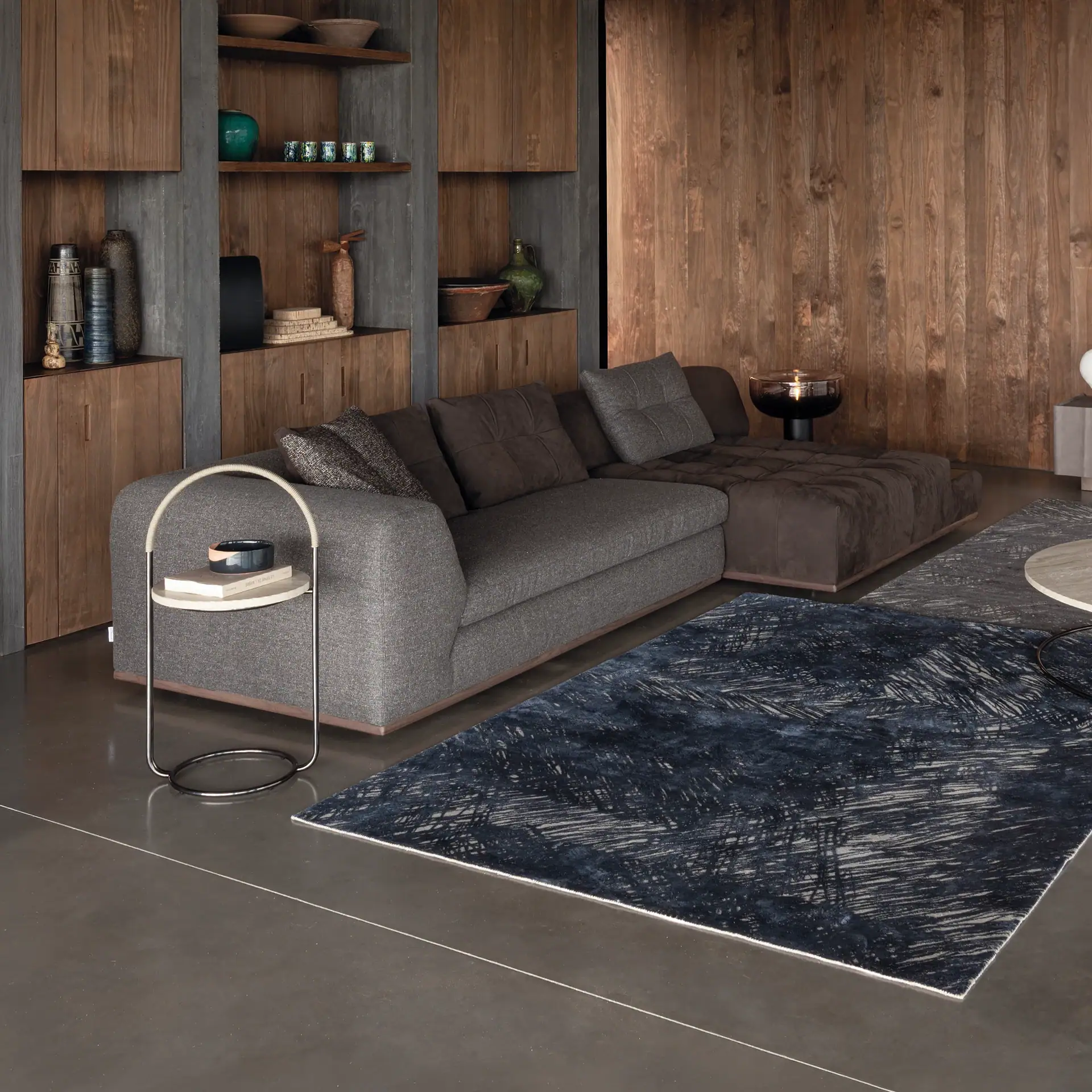 A modern living room with an L-shaped grey and brown sofa, abstract blue rugs, a side table, and built-in wooden shelving displaying decorative items.