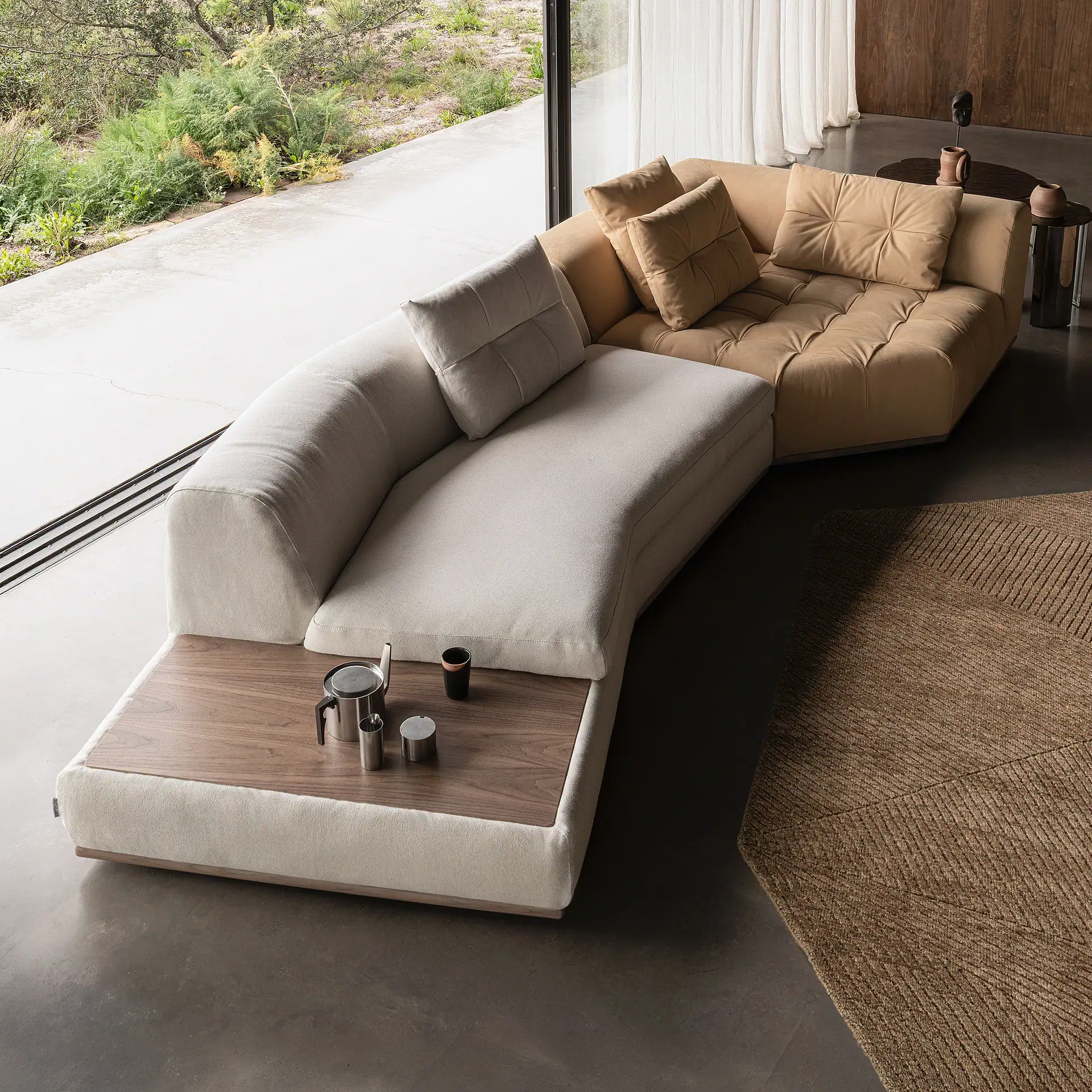 A curved sectional sofa in a modern living room. One section is light beige with a wooden end table holding coffee items, the other is camel-colored leather. Large windows overlook green foliage.