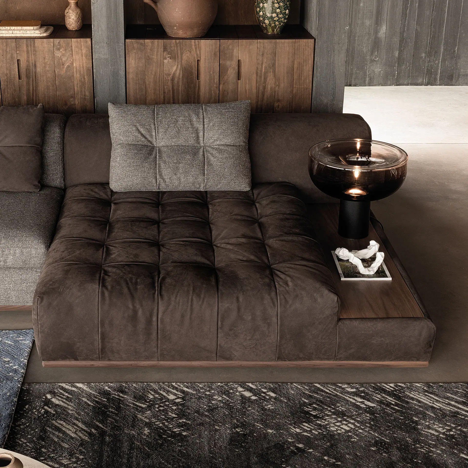 A dark brown tufted sectional sofa with a wooden base, integrated side table, and a modern lamp with a bowl-shaped shade. A grey pillow rests on the sofa, and a dark patterned rug covers the floor.