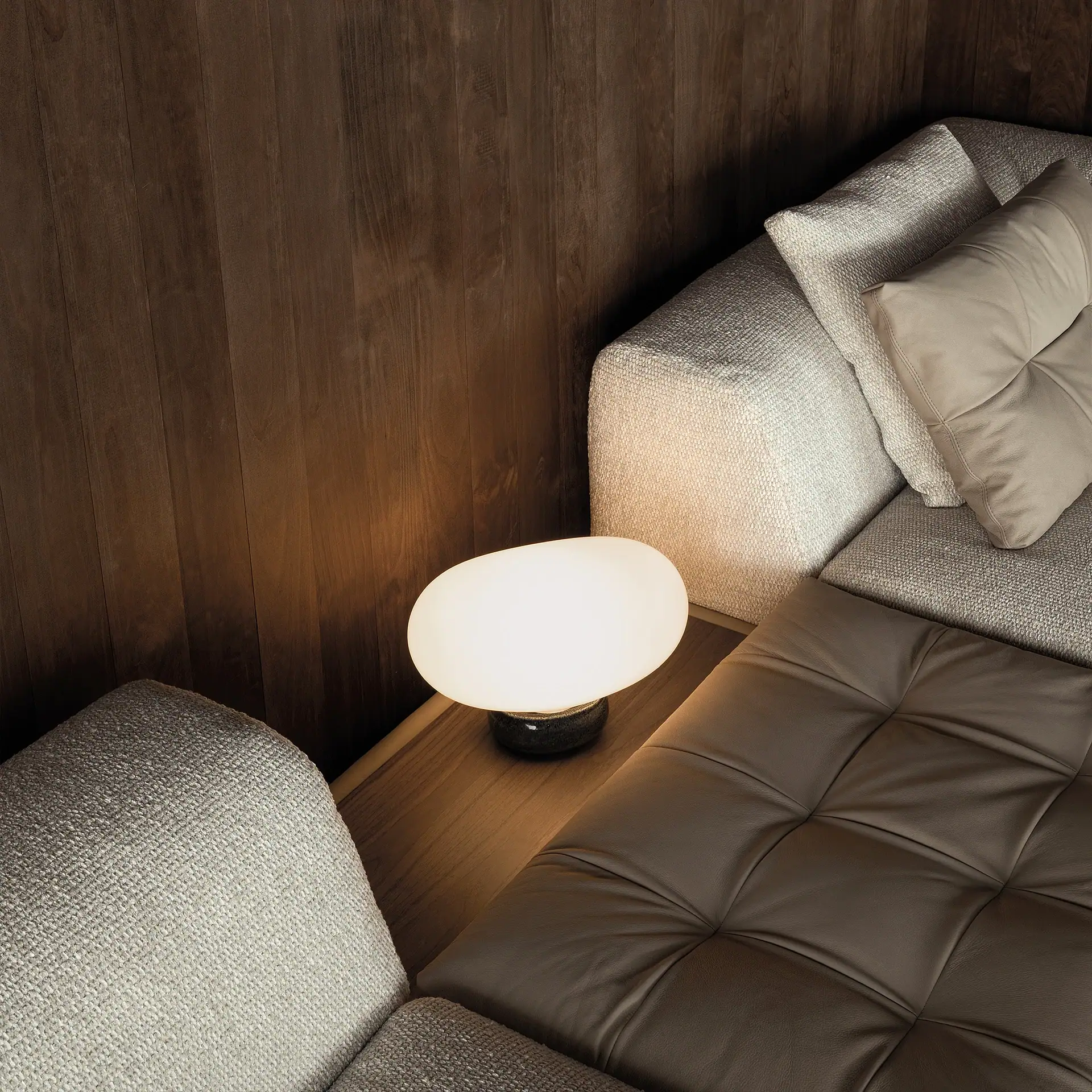 A close-up of a modern sofa corner with dark wood paneling, light fabric, brown leather cushions, and an oval-shaped glowing table lamp.