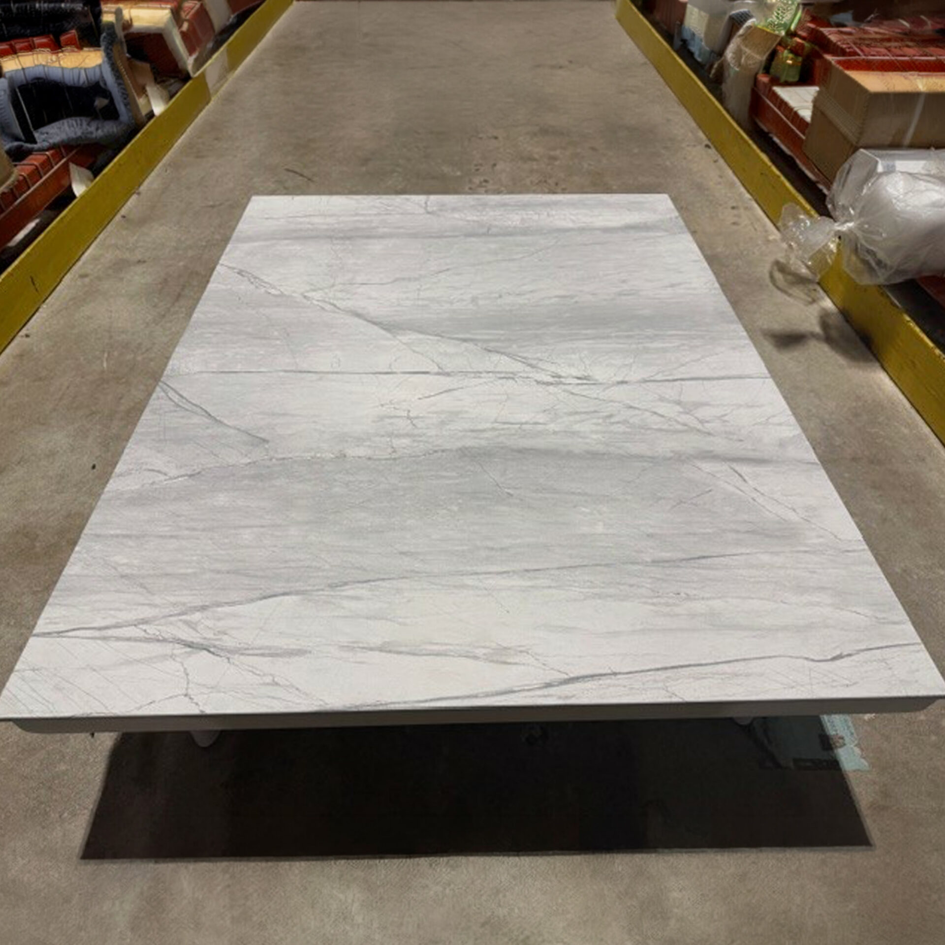 A rectangular light gray marble-patterned slab with dark veining, elevated above a concrete floor, flanked by yellow storage rails.