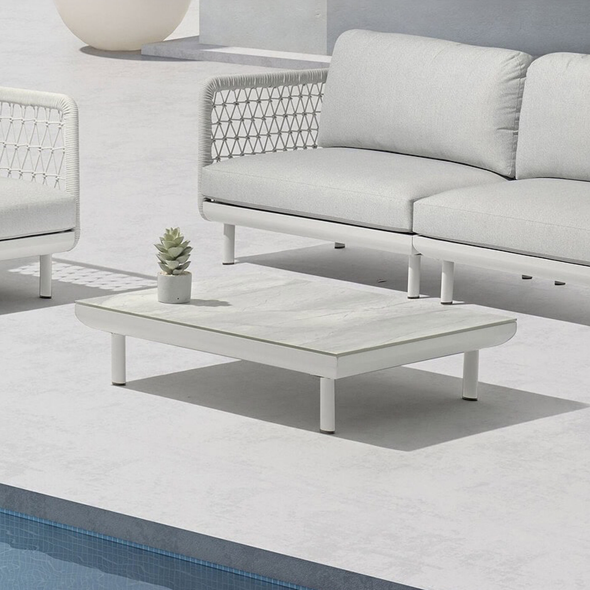 A modern outdoor patio scene with a light gray sofa, armchair, and a rectangular coffee table with a succulent, all in white frames, set on a light gray patio next to a blue swimming pool.