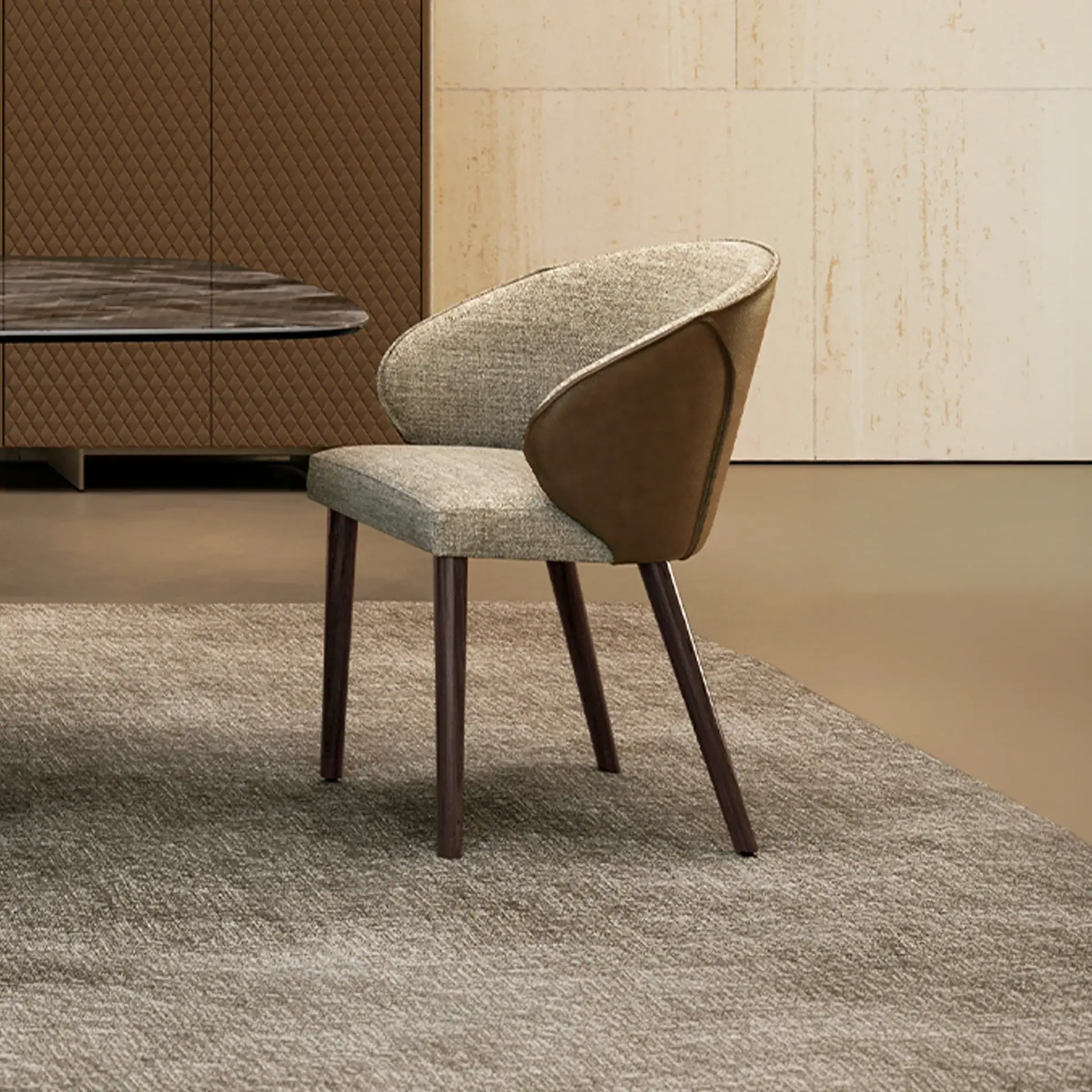 A modern dining chair with light gray fabric and dark brown leather accents, on a textured rug. A dark brown cabinet and light beige wall are in the background.