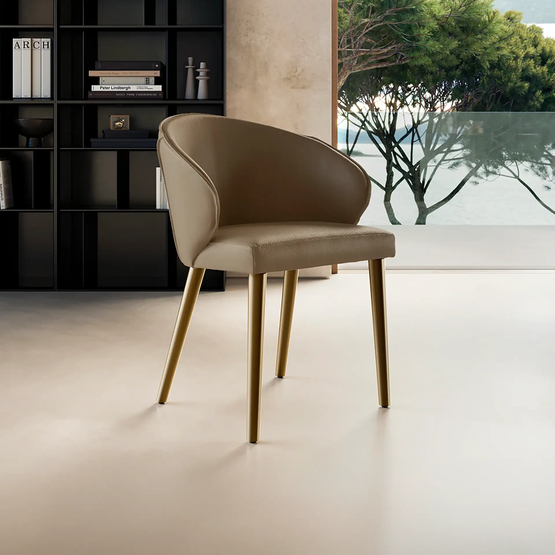A light brown upholstered chair with gold legs sits in a room with a black bookshelf and a large window overlooking green trees and water.