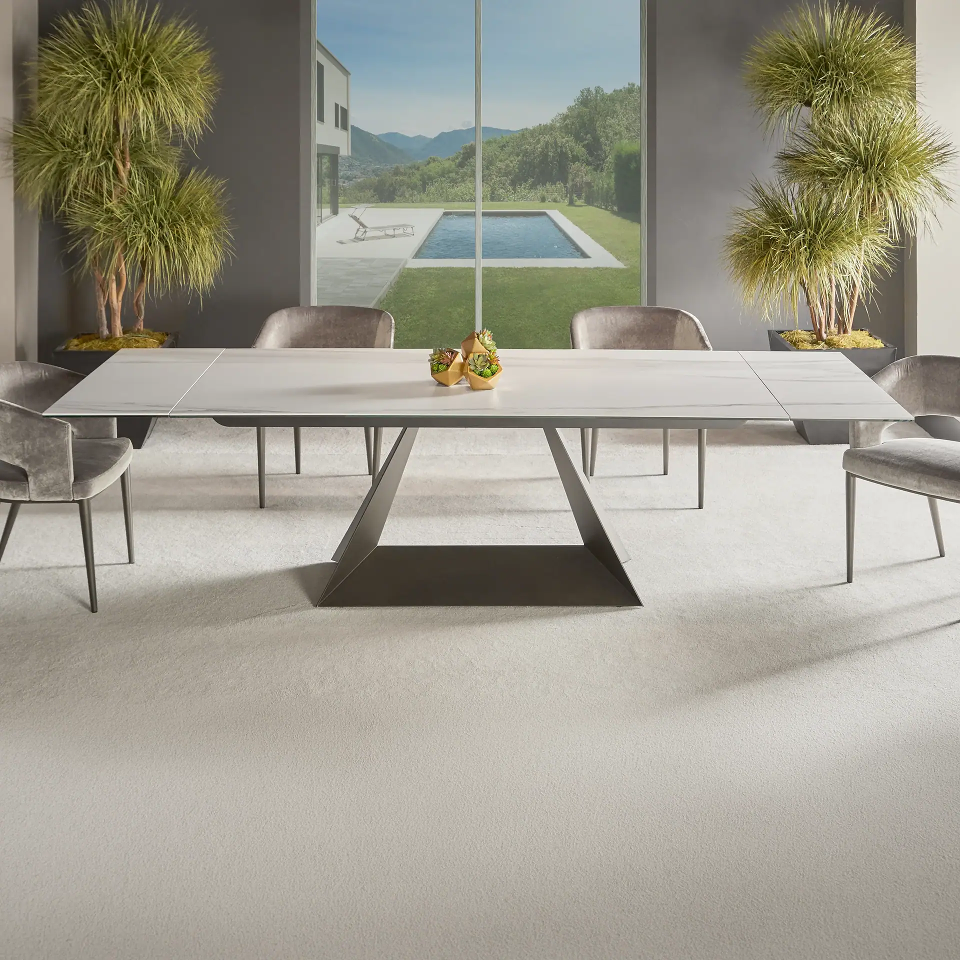 A modern dining room with a marble-effect table, grey velvet chairs, and a large window overlooking a house, swimming pool, and mountains.