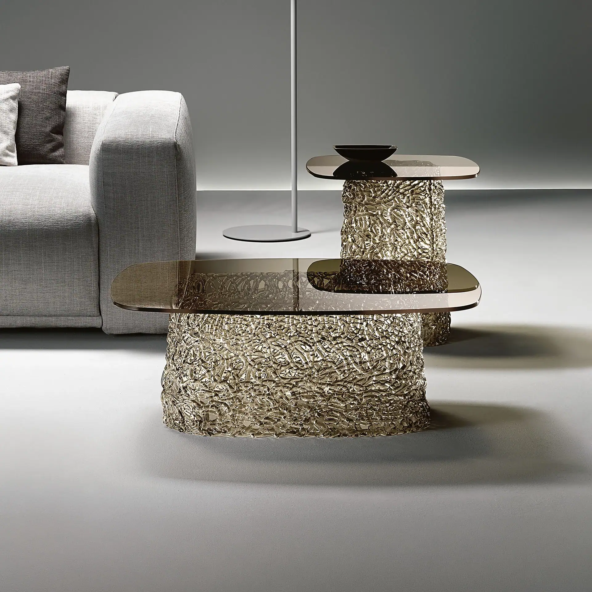 Two modern coffee tables with textured gold bases and dark glass tops, next to a light grey sofa with pillows in a minimalist room.