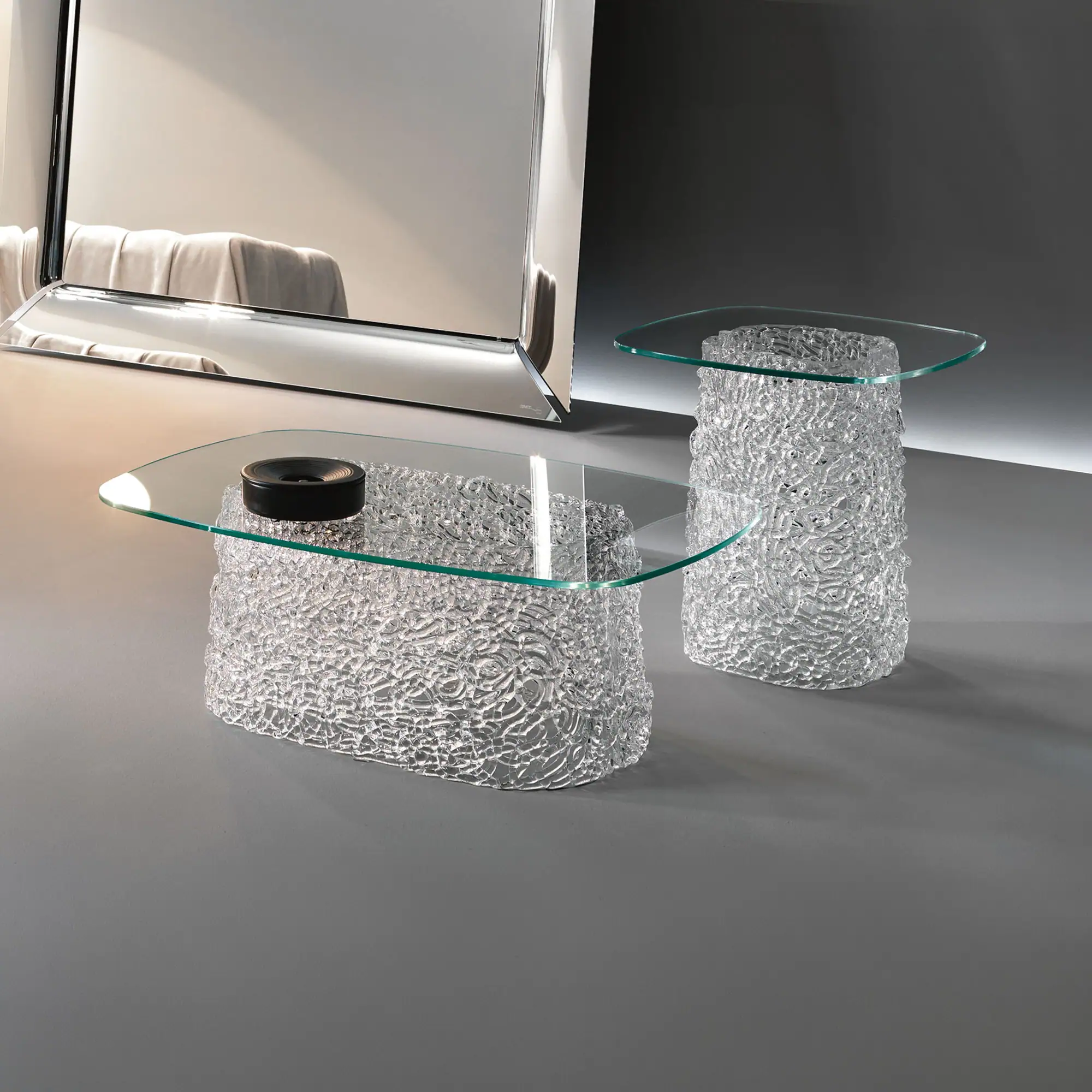 Two modern glass tables with clear, textured bases and glass tops. A large mirror stands on the left, reflecting a light-colored fabric.