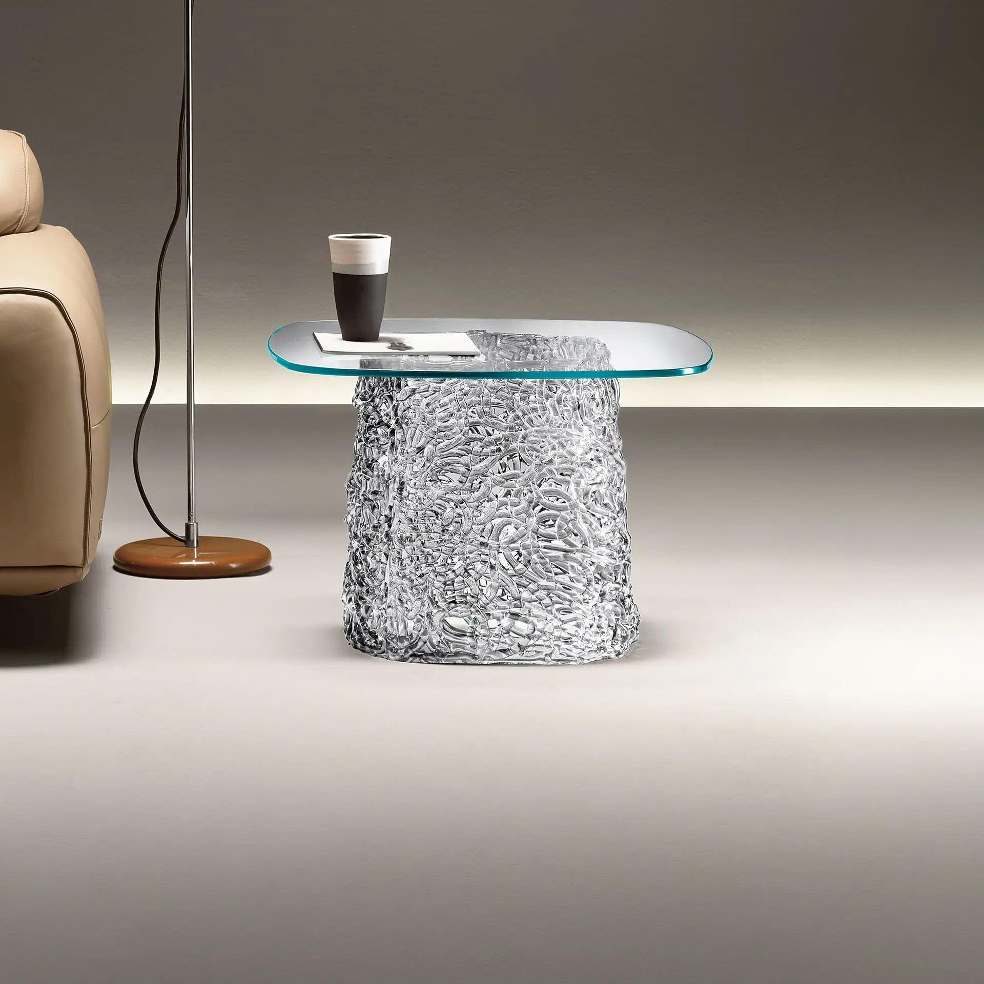 A modern side table with an oval glass top and a cylindrical, textured clear base, next to a light brown leather sofa and a floor lamp.