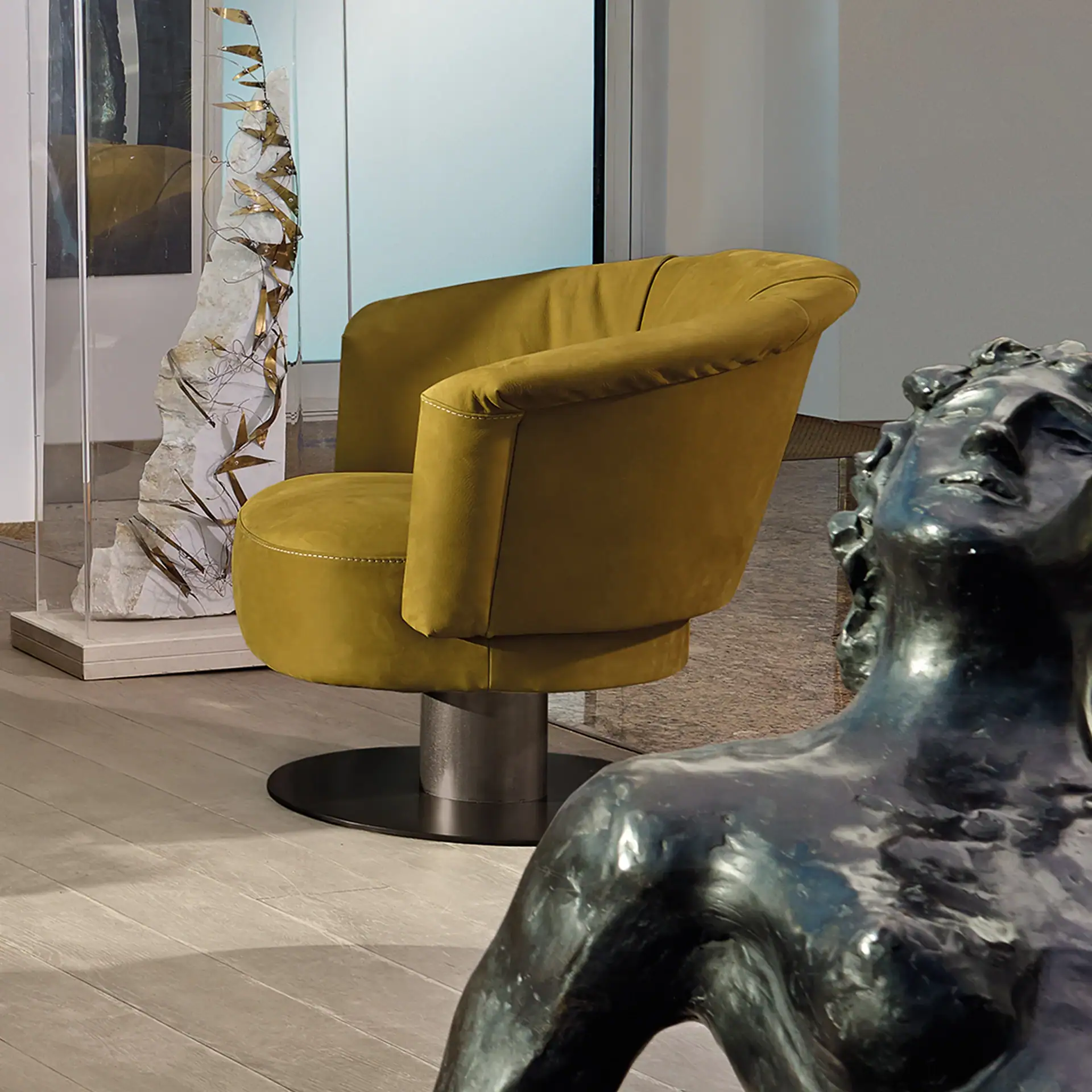Mustard yellow armchair on a dark base, flanked by a metallic leaf sculpture in acrylic and a bronze human torso sculpture, on a light wooden floor.