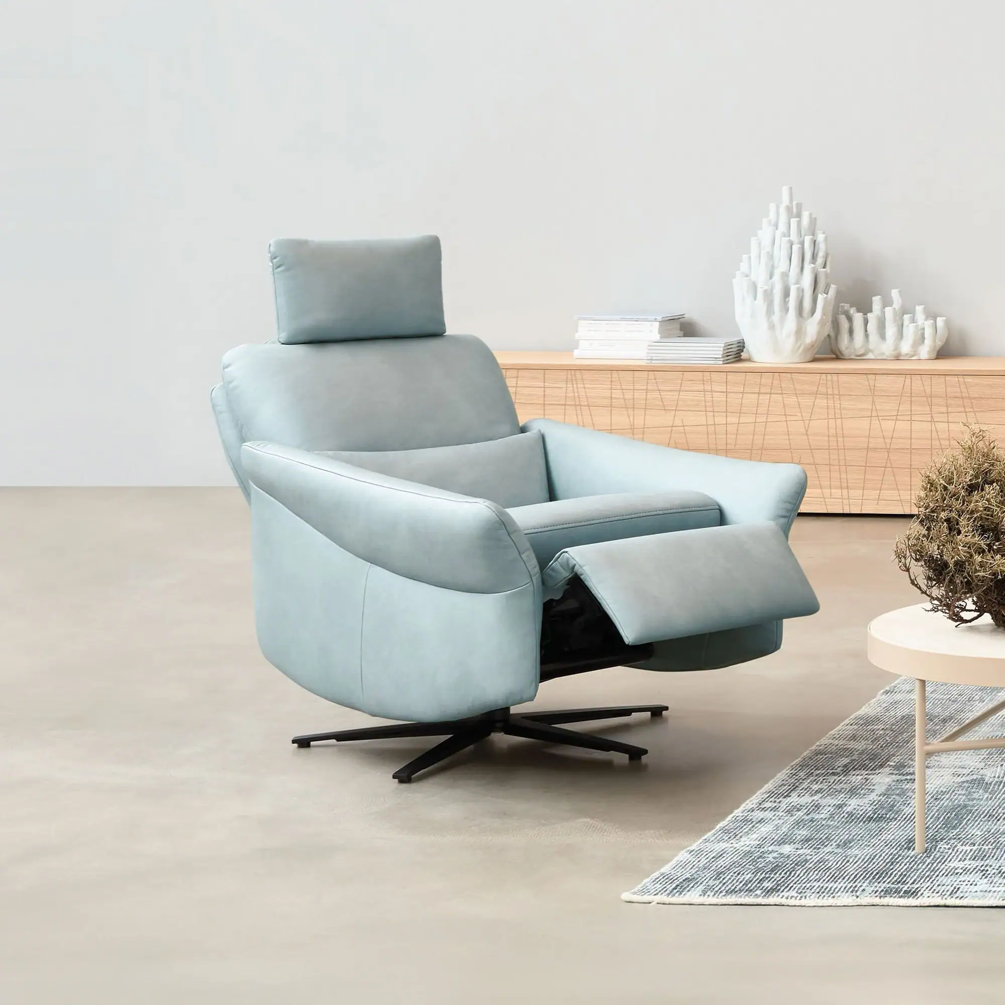 A light blue recliner chair with an extended footrest sits on a gray floor. In the background, a wooden credenza holds white books and coral-like decorations. A round coffee table with a dried plant and a patterned rug are in the foreground.