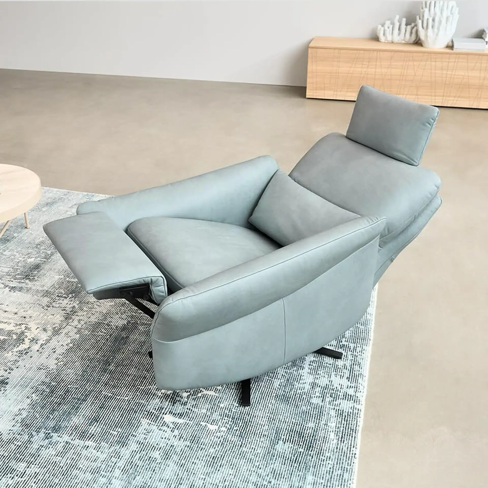 A light blue-grey recliner chair, partially reclined with an extended footrest, sits on a patterned rug in a modern room. A wooden cabinet with white decor is in the background.