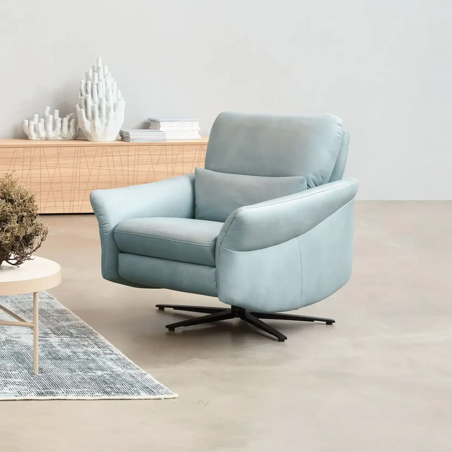 A light blue upholstered armchair with a black swivel base sits on a light gray floor. A light wood credenza with white coral-like decor and books is in the background. A round side table with a dried plant and a gray rug are to the left.