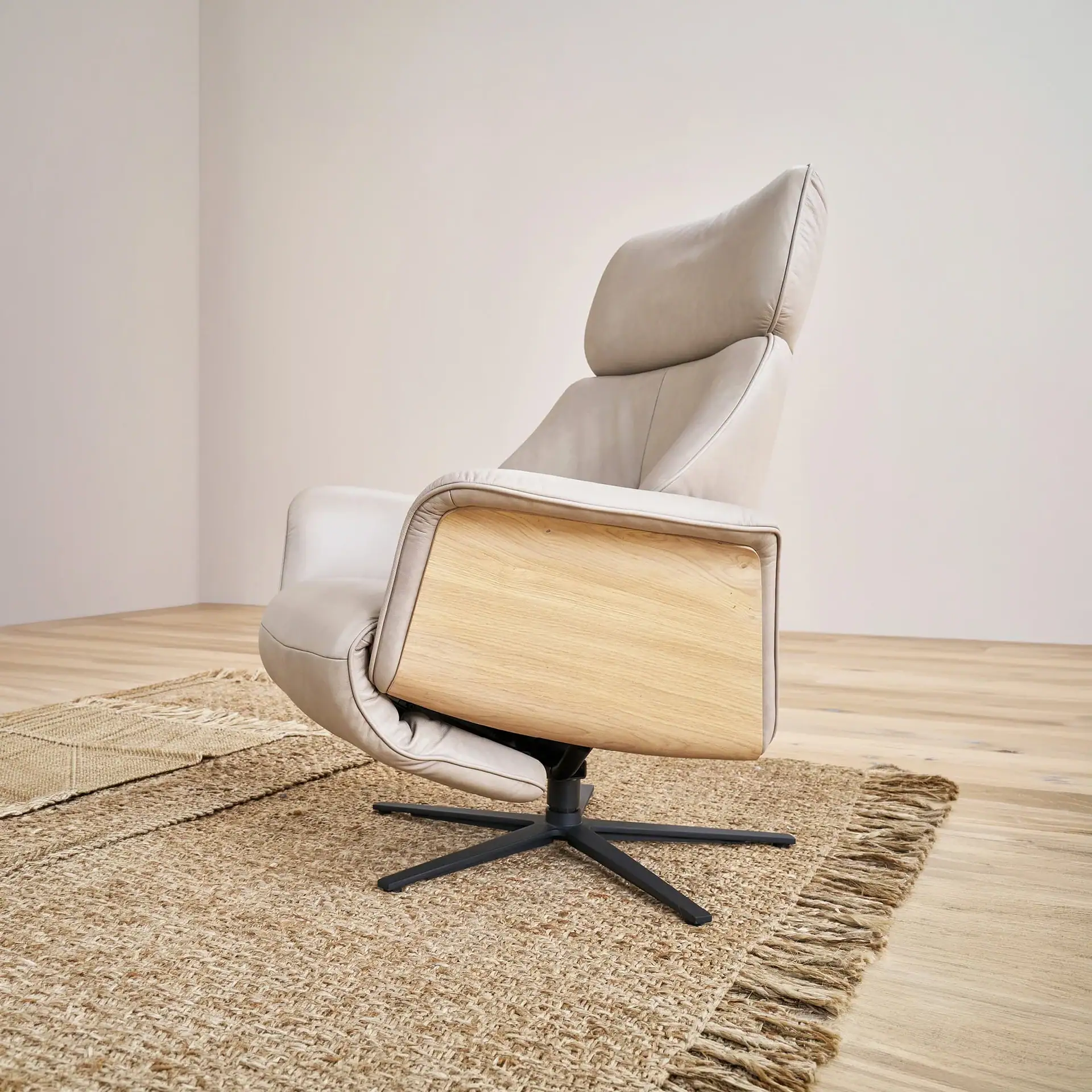 A light gray leather recliner chair with wooden armrests sits on a natural jute rug on a wooden floor.