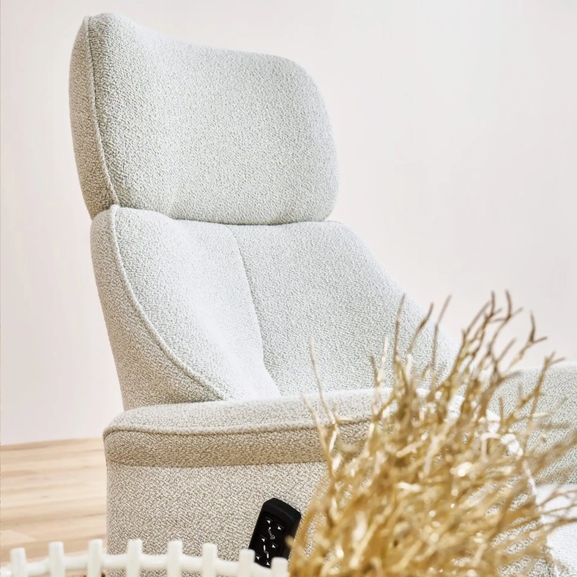 A light-colored, textured armchair with a high back, a remote control on its seat, and golden decorative branches in the foreground.
