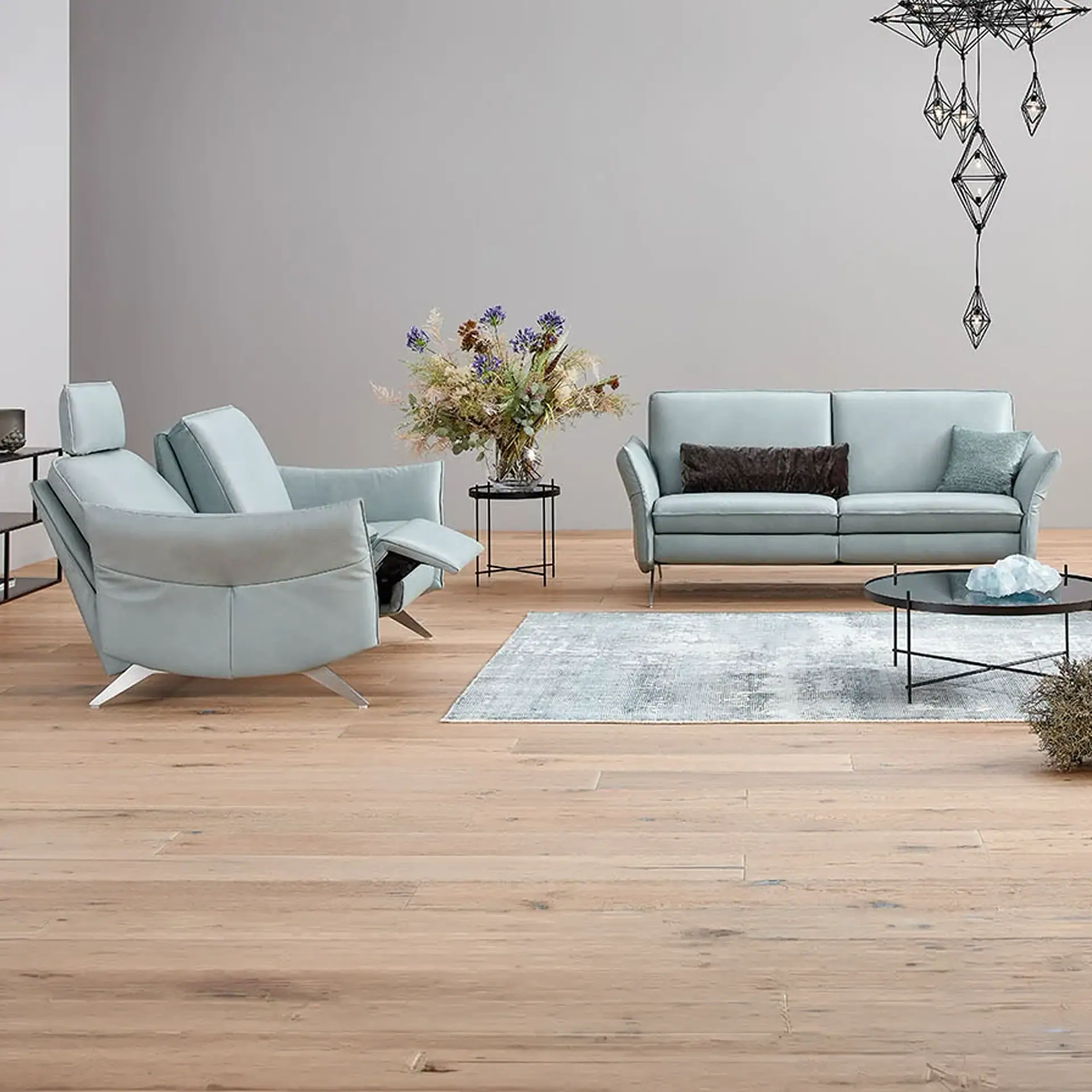 A modern living room with light blue-grey sofa and recliner, a light blue and white rug, a black coffee table, and a geometric chandelier.