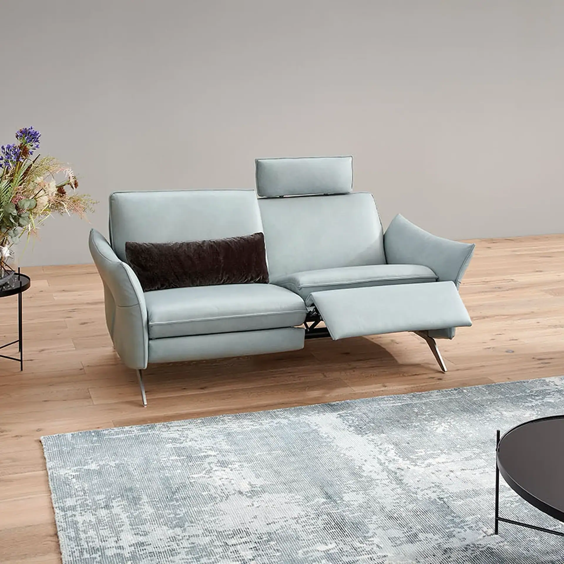 A light blue two-seater recliner sofa with one seat extended, a raised headrest, and a dark brown cushion, set on a wooden floor with a patterned rug and a floral arrangement on a side table.