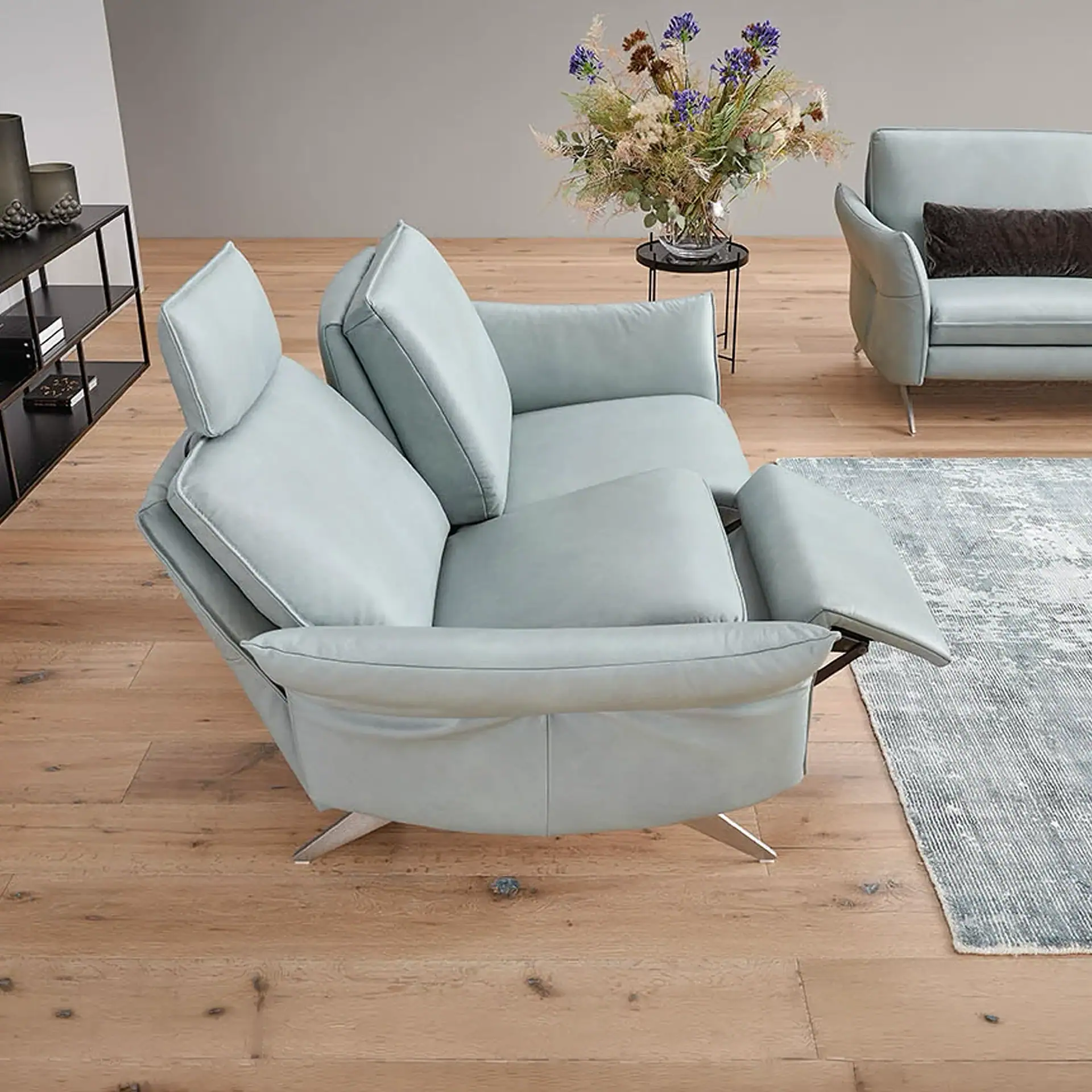 A light blue recliner sofa with an extended footrest and adjustable headrest in a modern living room. A matching sofa, floral arrangement, and shelving unit are in the background.