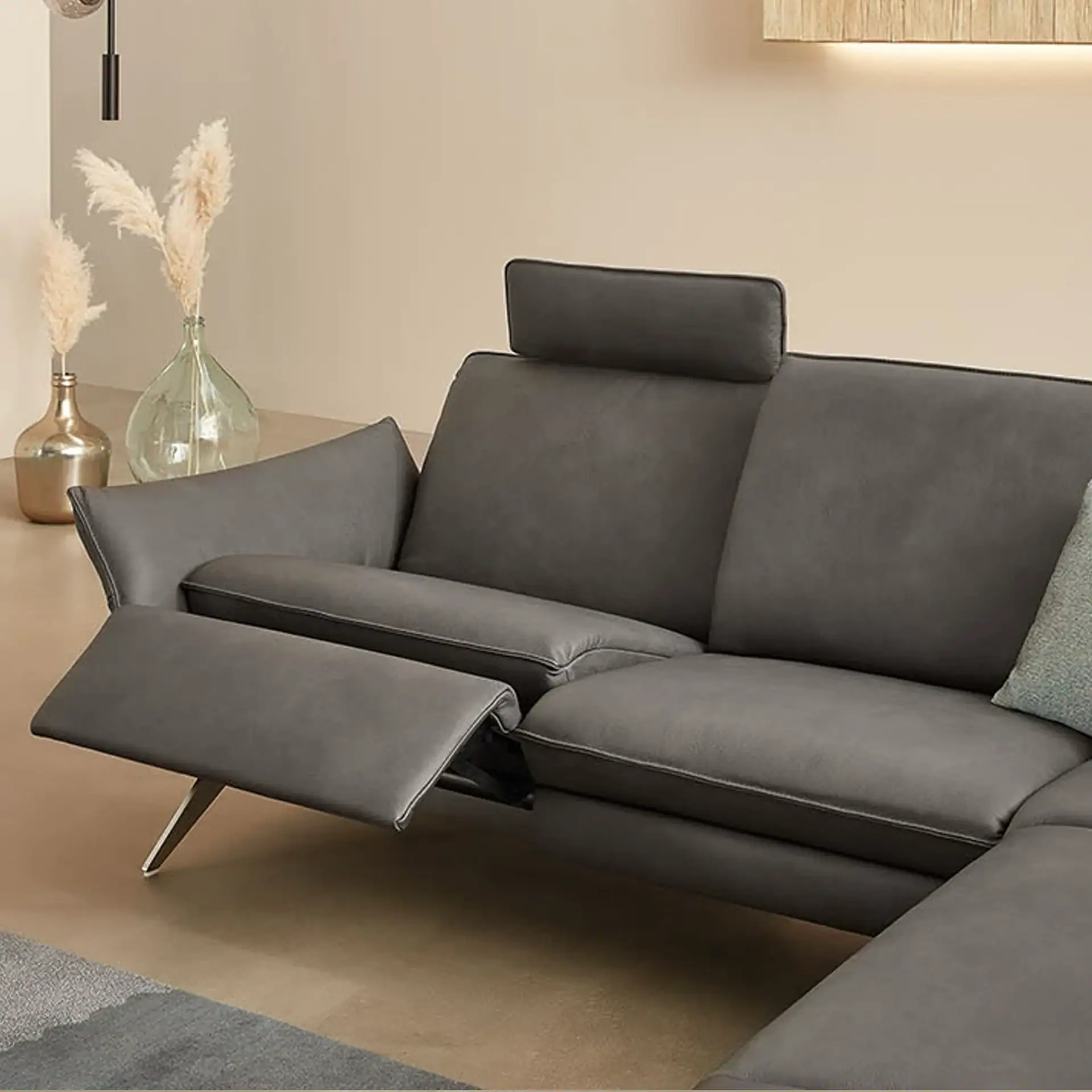 A dark grey sectional sofa with an extended recliner footrest and an adjustable headrest. Pampas grass in vases and a wooden wall light are in the background.