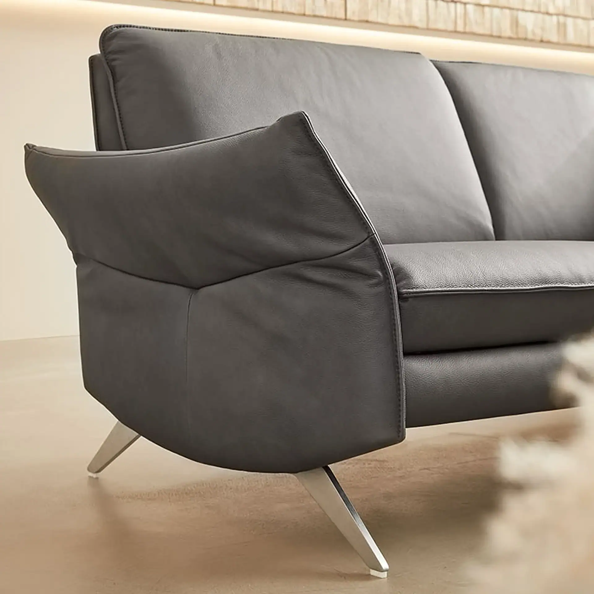 A close-up of a modern dark gray leather sofa with curved armrests and silver metal legs, set against a textured, illuminated wall.