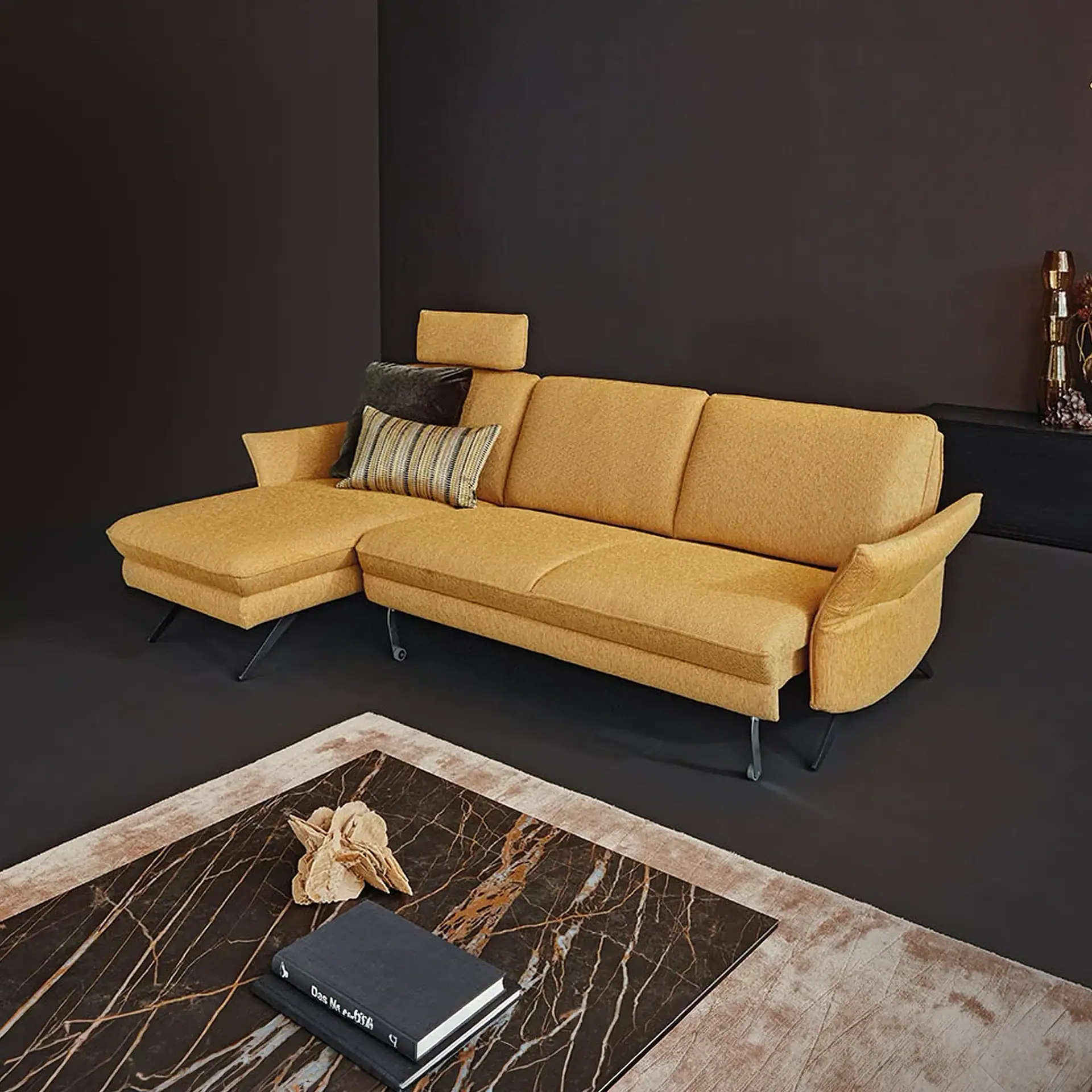 Mustard yellow L-shaped sectional sofa with dark metal legs in a modern room. A marble coffee table with books and a decorative object sits on a light rug in the foreground.
