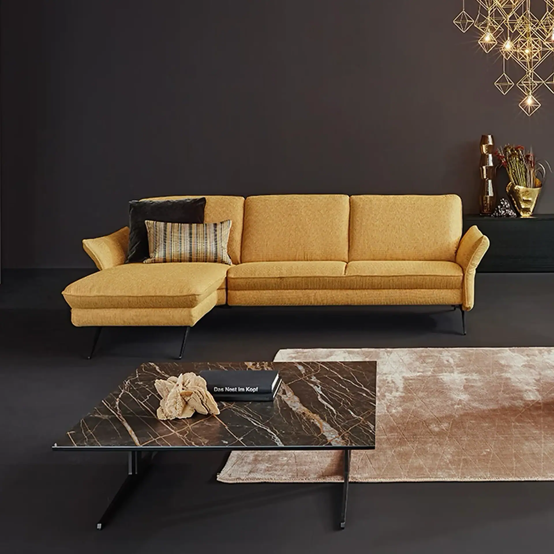 A modern living room featuring a mustard yellow L-shaped sectional sofa, a dark marble coffee table, and a geometric gold chandelier against a dark gray wall.