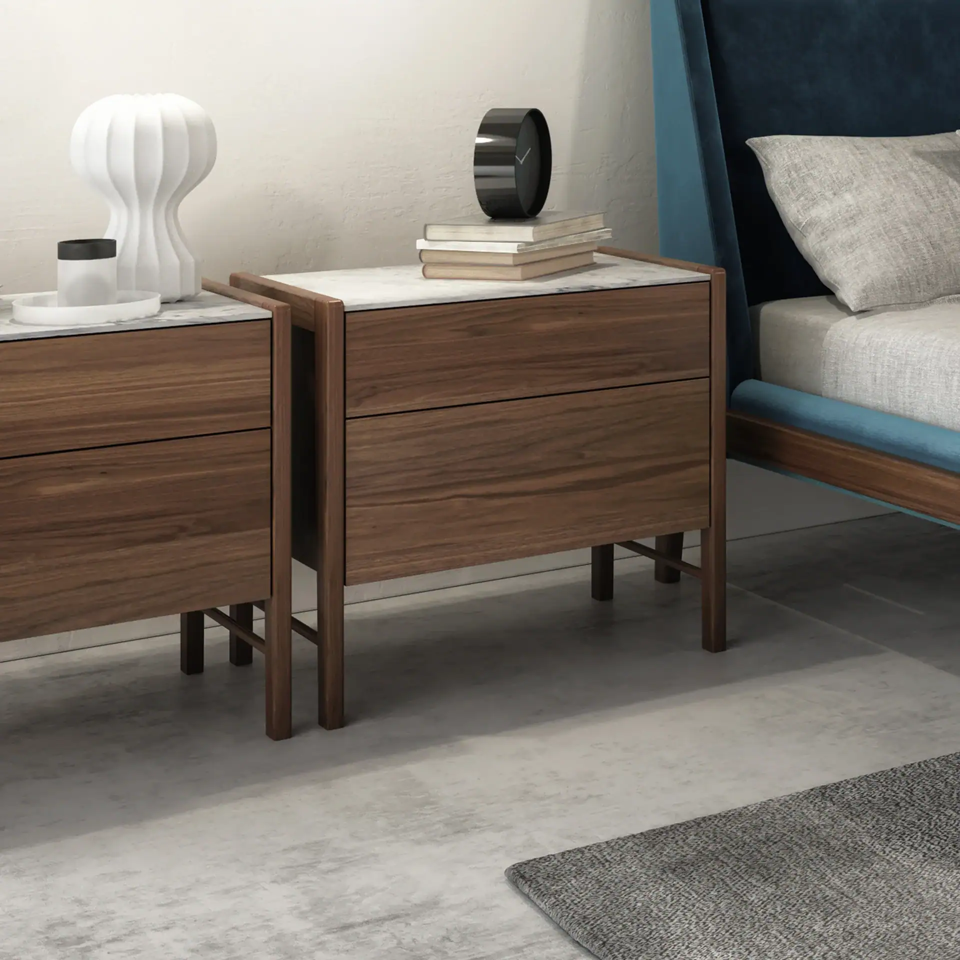 Two modern wooden nightstands with marble tops next to a bed with a blue velvet headboard. One nightstand has a white vase and tray, the other has books and a black clock.