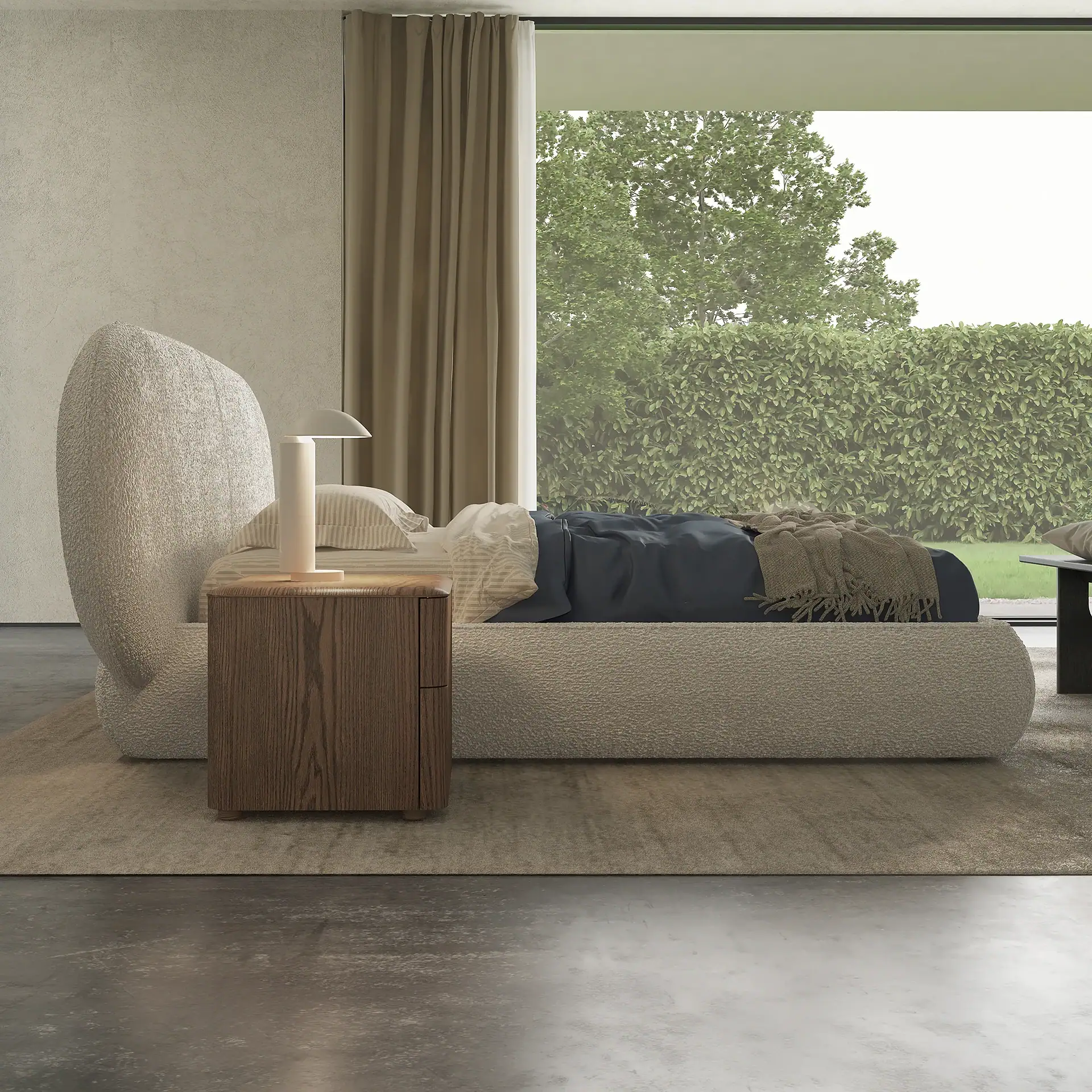 A modern bedroom with a large, light beige textured bed, a wooden nightstand, and a white lamp, facing a floor-to-ceiling window overlooking green trees and a hedge.
