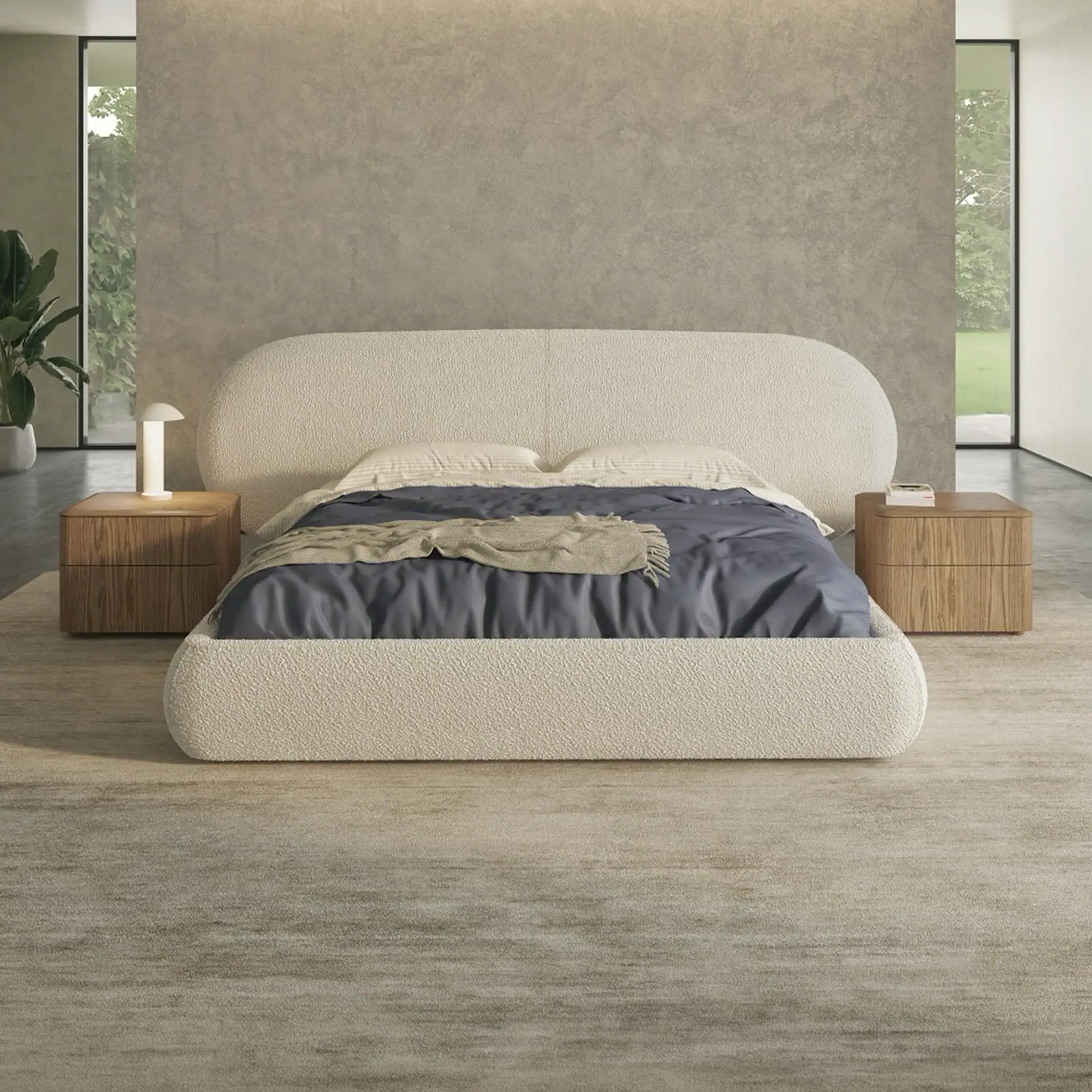 A modern bedroom with a cream-colored, rounded bed, dark blue bedding, wooden nightstands, and large windows revealing green foliage.
