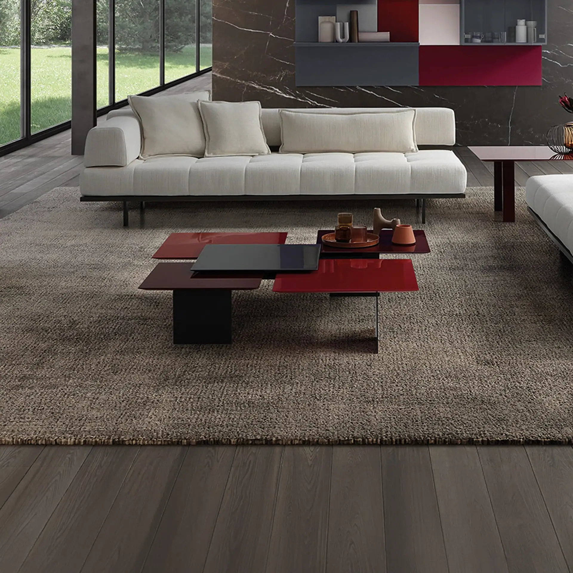 A modern living room with a light-colored sofa, a multi-level coffee table with red and dark gray tops, and a dark marble wall with colorful shelves. Large windows show a green lawn outside.
