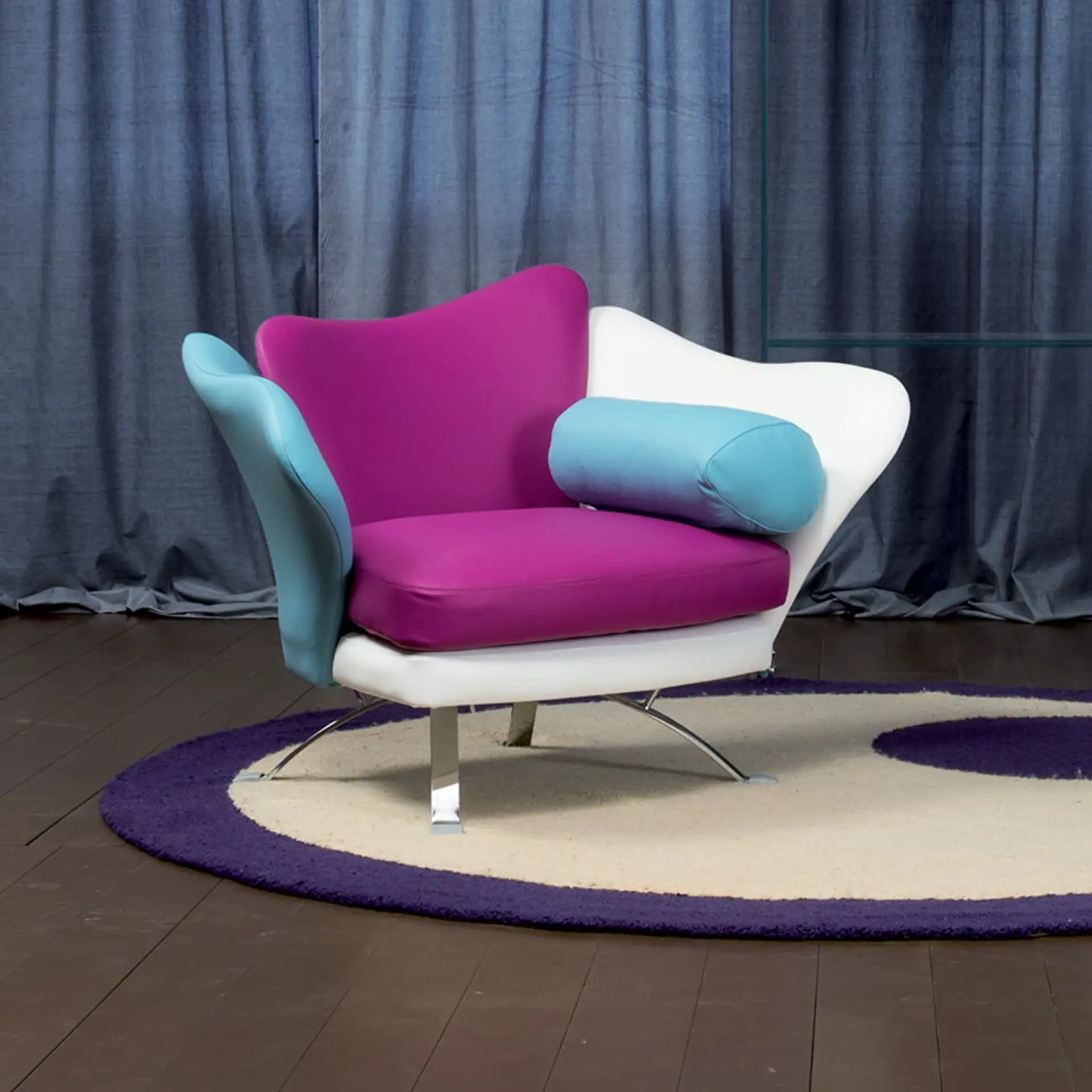 A modern armchair with magenta, white, and light blue upholstery, chrome legs, sitting on a round cream and purple rug on a dark wood floor, with dark blue curtains in the background.