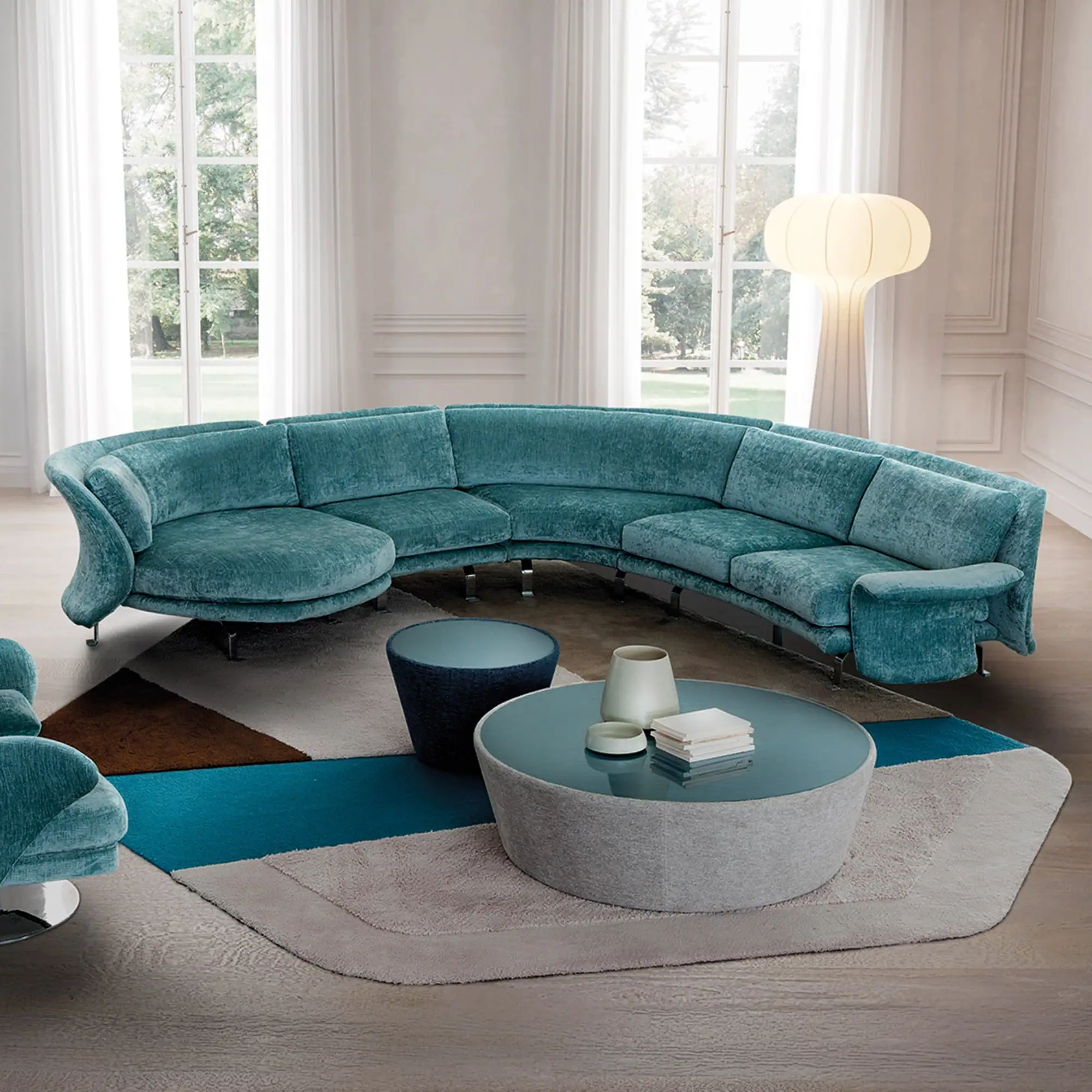 A luxurious living room with a large, curved teal sectional sofa, a round glass coffee table, and a geometric rug. Two tall windows overlook a garden, and a modern floor lamp stands near a white fireplace.