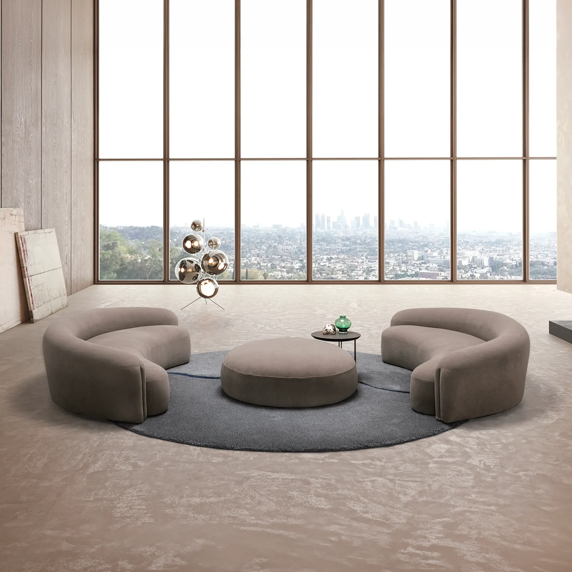 A modern living room with two curved taupe sofas and a round ottoman on a grey rug, facing a large window with a city view. A spherical floor lamp stands on the left.