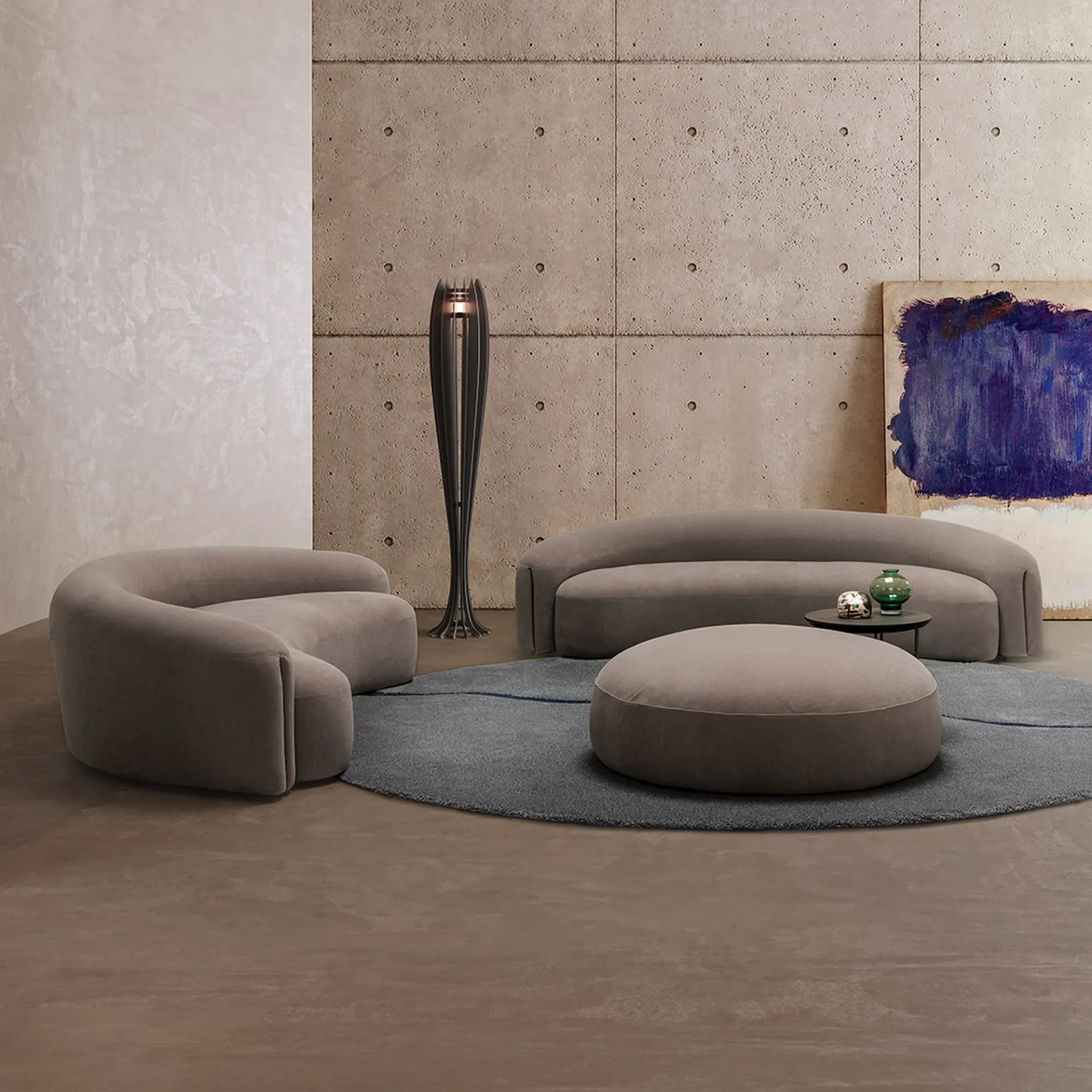 A modern living room with two curved taupe sofas, a round ottoman, and a dark grey rug. The room features a concrete wall, a textured light wall, a sculptural floor lamp, and an abstract painting.