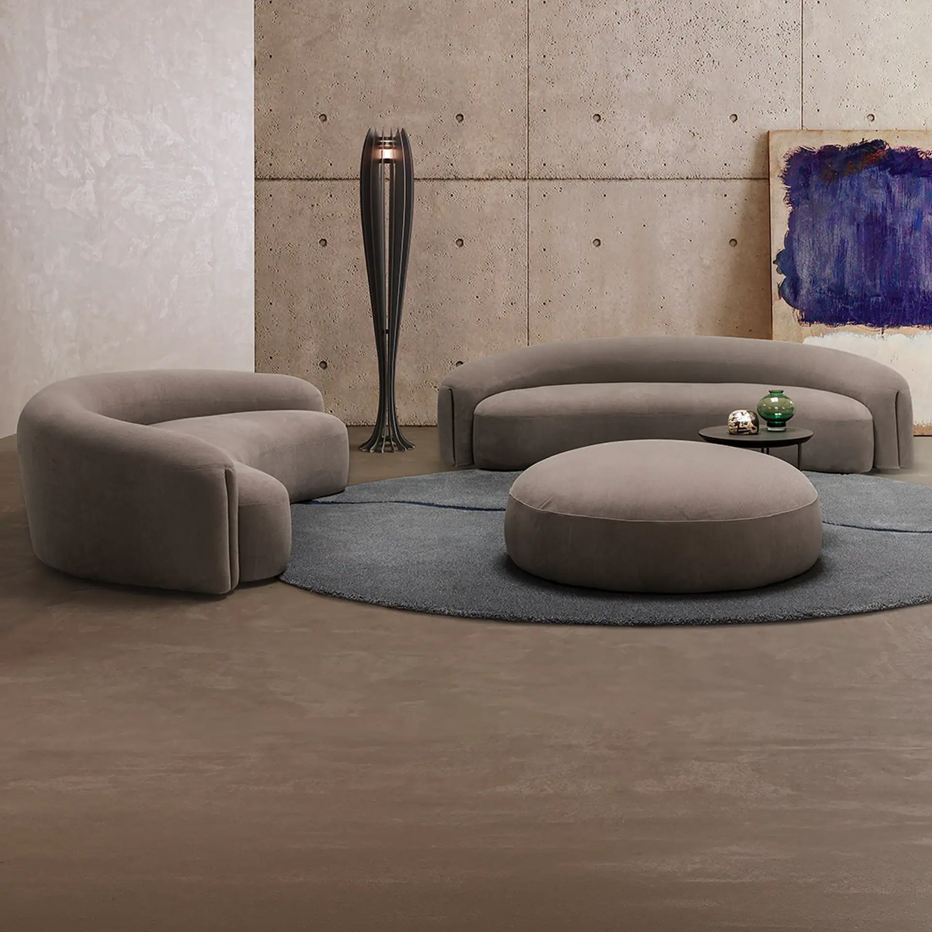 A modern living room with curved taupe sofa, armchair, and ottoman on a gray rug, a sculptural floor lamp, and an abstract blue painting against concrete and textured walls.