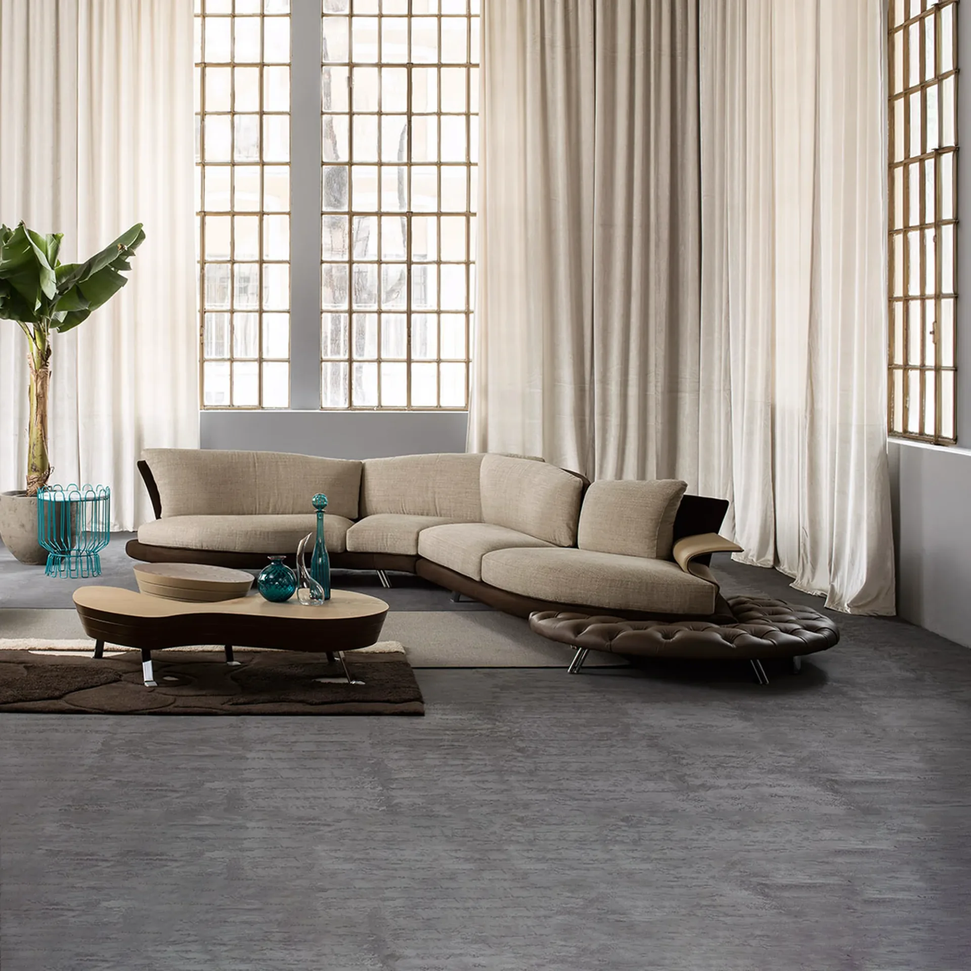A modern living room with a large, curved, light beige sectional sofa, a kidney-bean shaped coffee table, and industrial windows with sheer curtains.