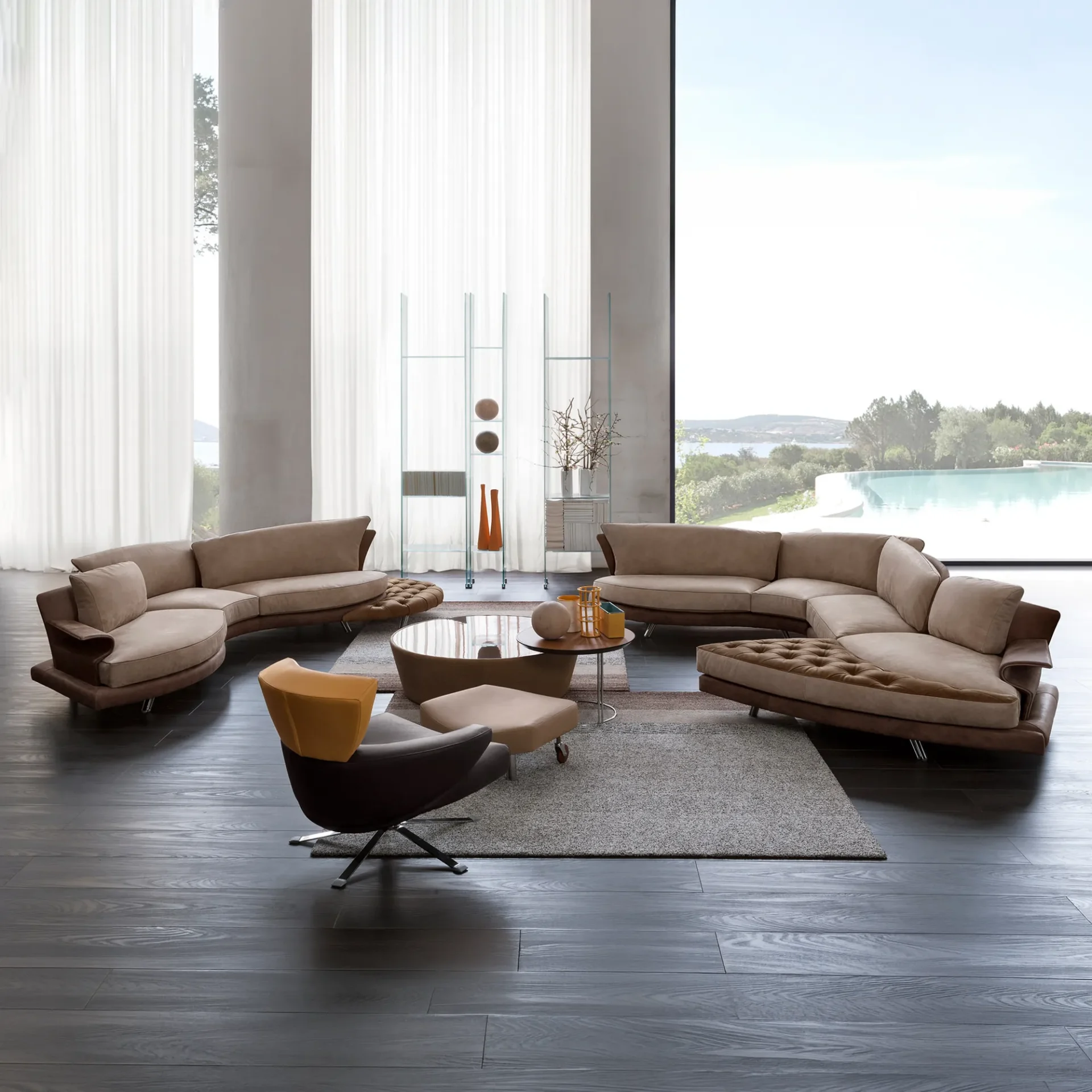 A modern living room with two curved beige sofas, a glass coffee table, and an armchair, set against large windows overlooking a swimming pool and a scenic bay.