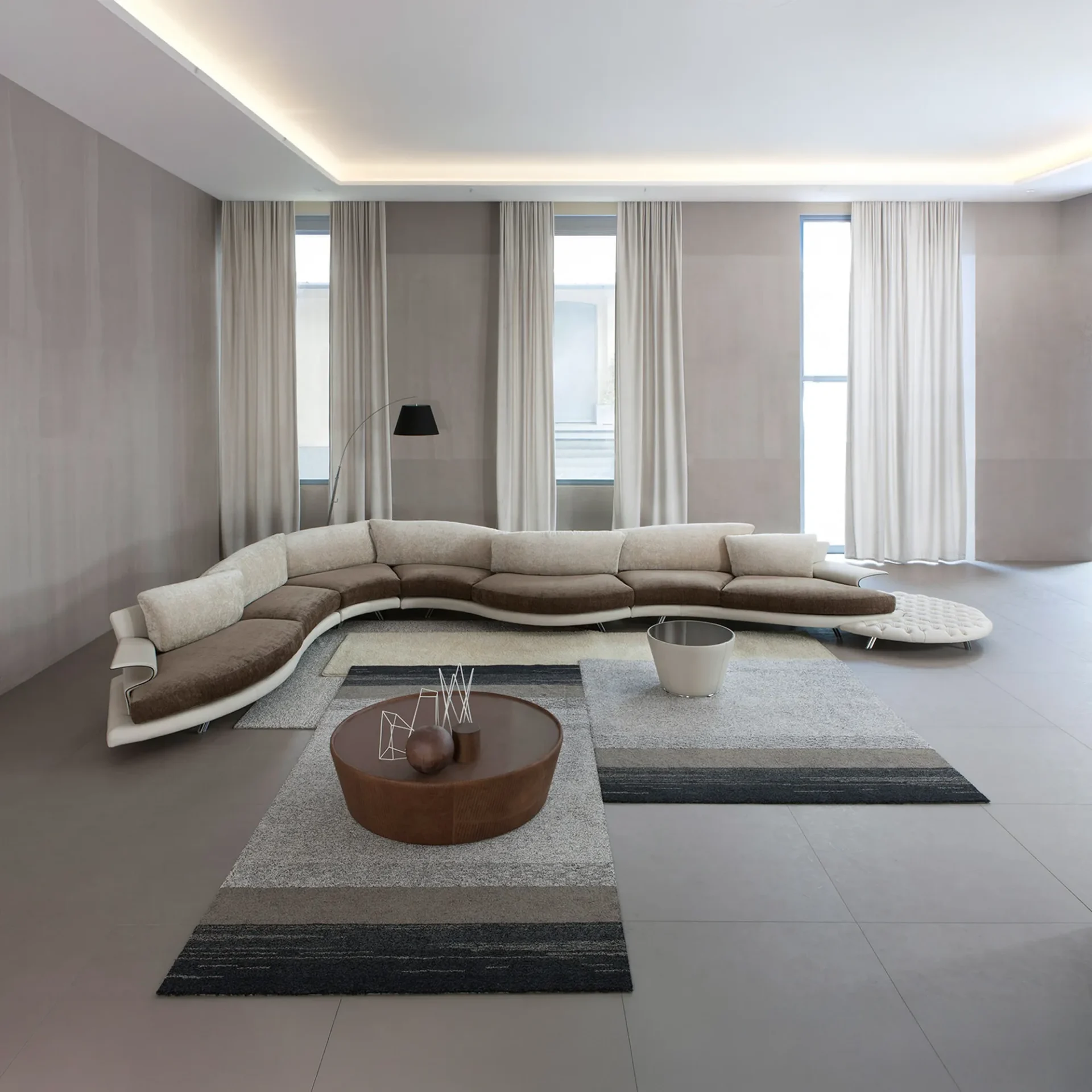 A modern living room with a large, curved brown and beige sofa, two striped rugs, a round coffee table, and tall windows with drapes.