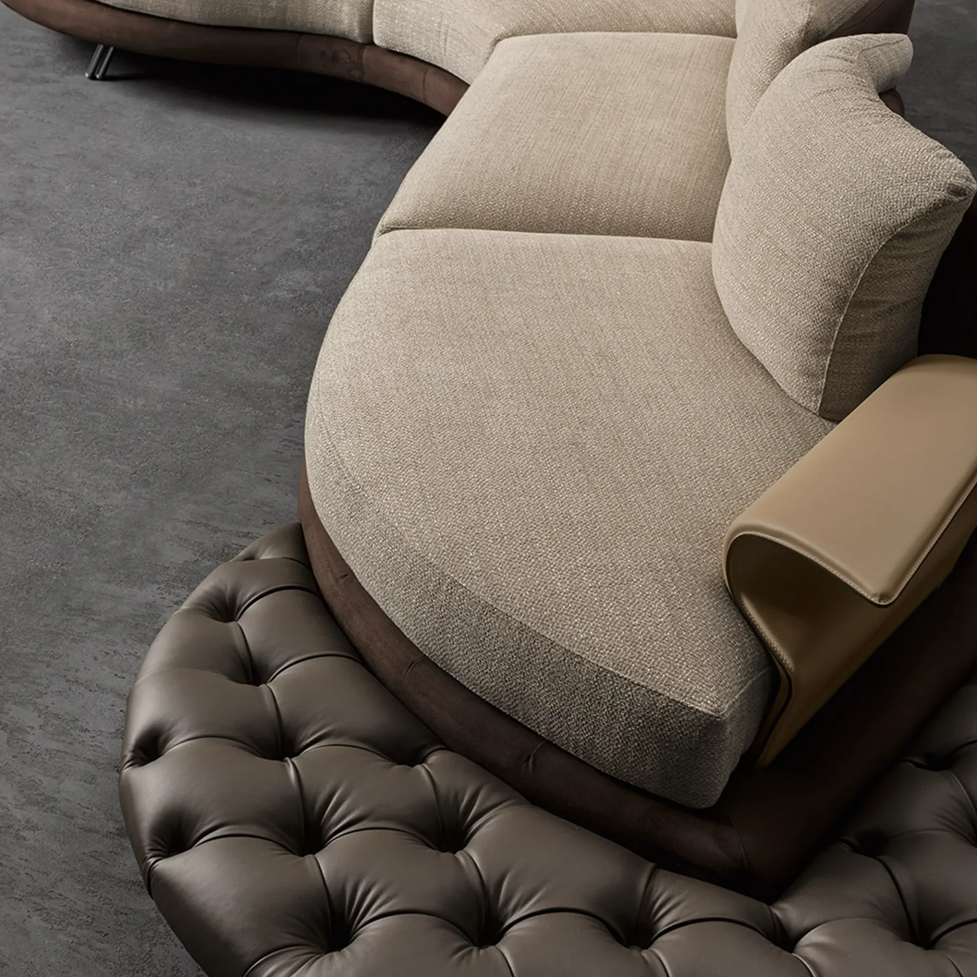 A curved sofa with light beige fabric cushions, dark brown base, and a matching tufted leather ottoman on a dark gray floor.