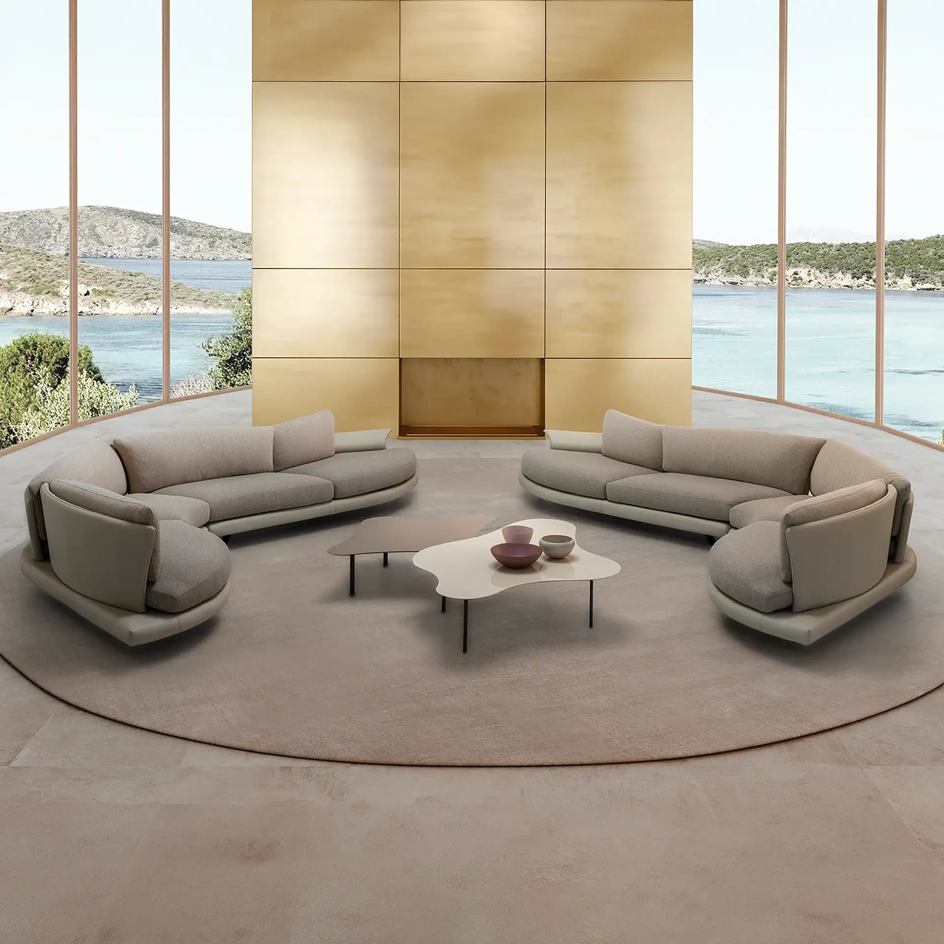 A luxurious living room with two Super Roy Modular Curved Sofas, two organic coffee tables, and a circular rug, set against a panoramic window view of a blue bay and green hills, with a golden panel wall.