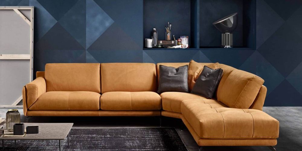 An L-shaped tan leather sectional sofa with dark grey pillows in a modern living room with a geometric blue wall, a coffee table, and art supplies. Glamour Modular Sectional Sofa by Bracci