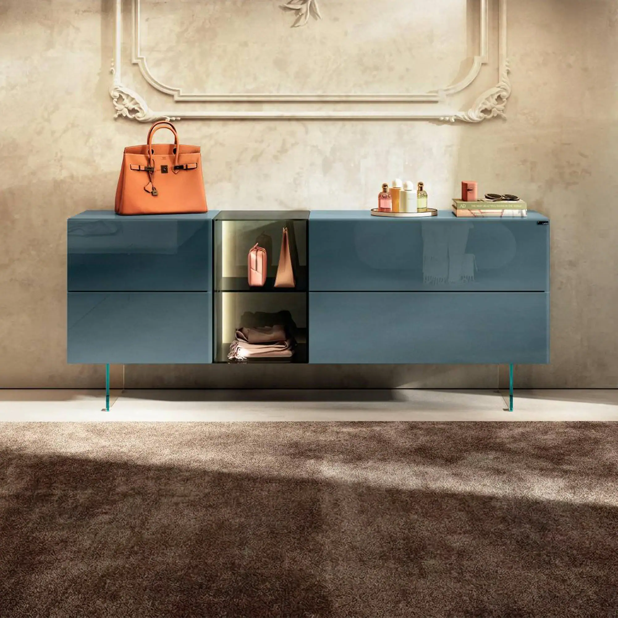 A glossy blue-grey sideboard with glass legs, an orange handbag, perfume bottles, and books, set against a textured beige wall with ornate molding.