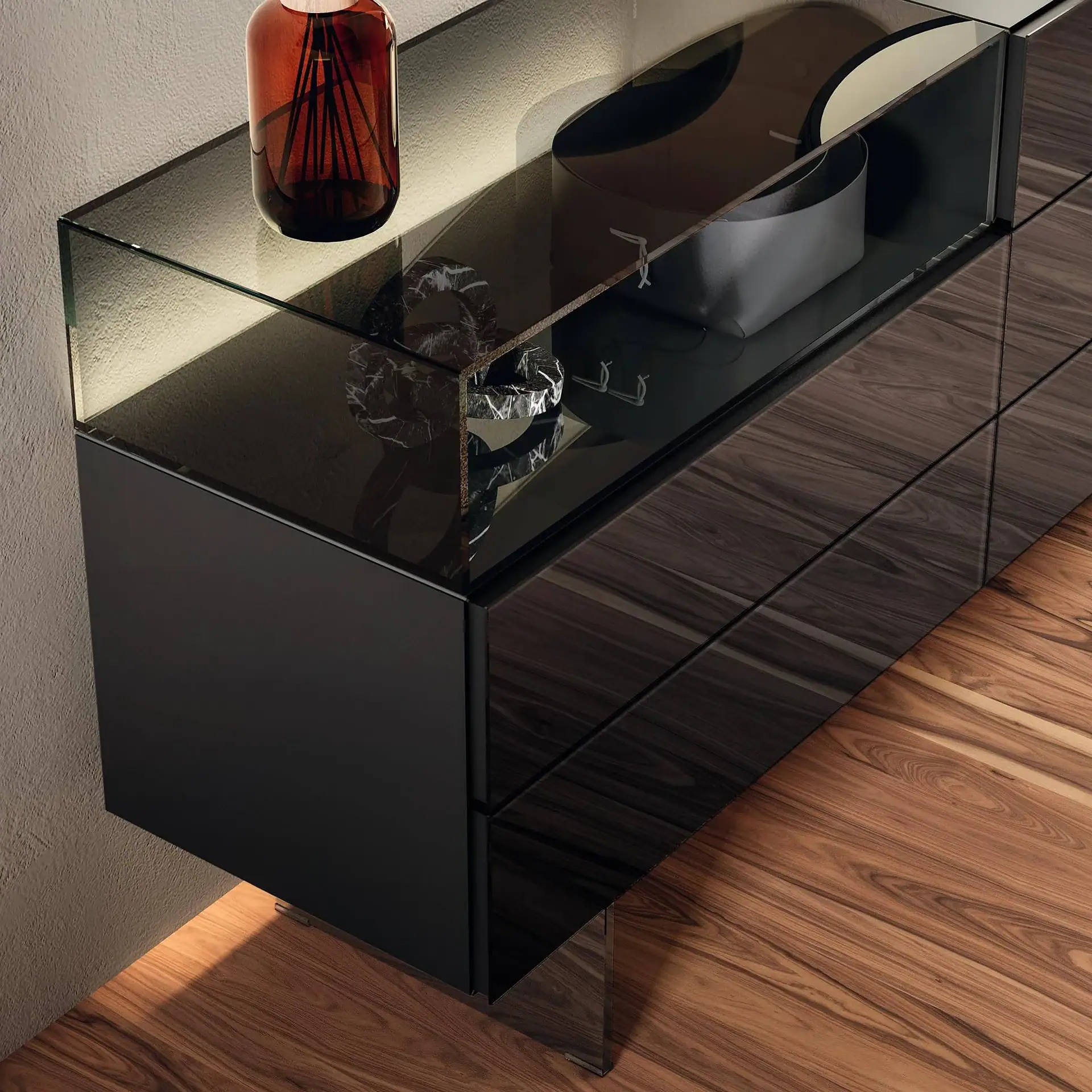A modern dark wood veneer sideboard with a clear glass top, an illuminated smoked glass display shelf, and decorative objects, set against a textured wall and wooden floor.