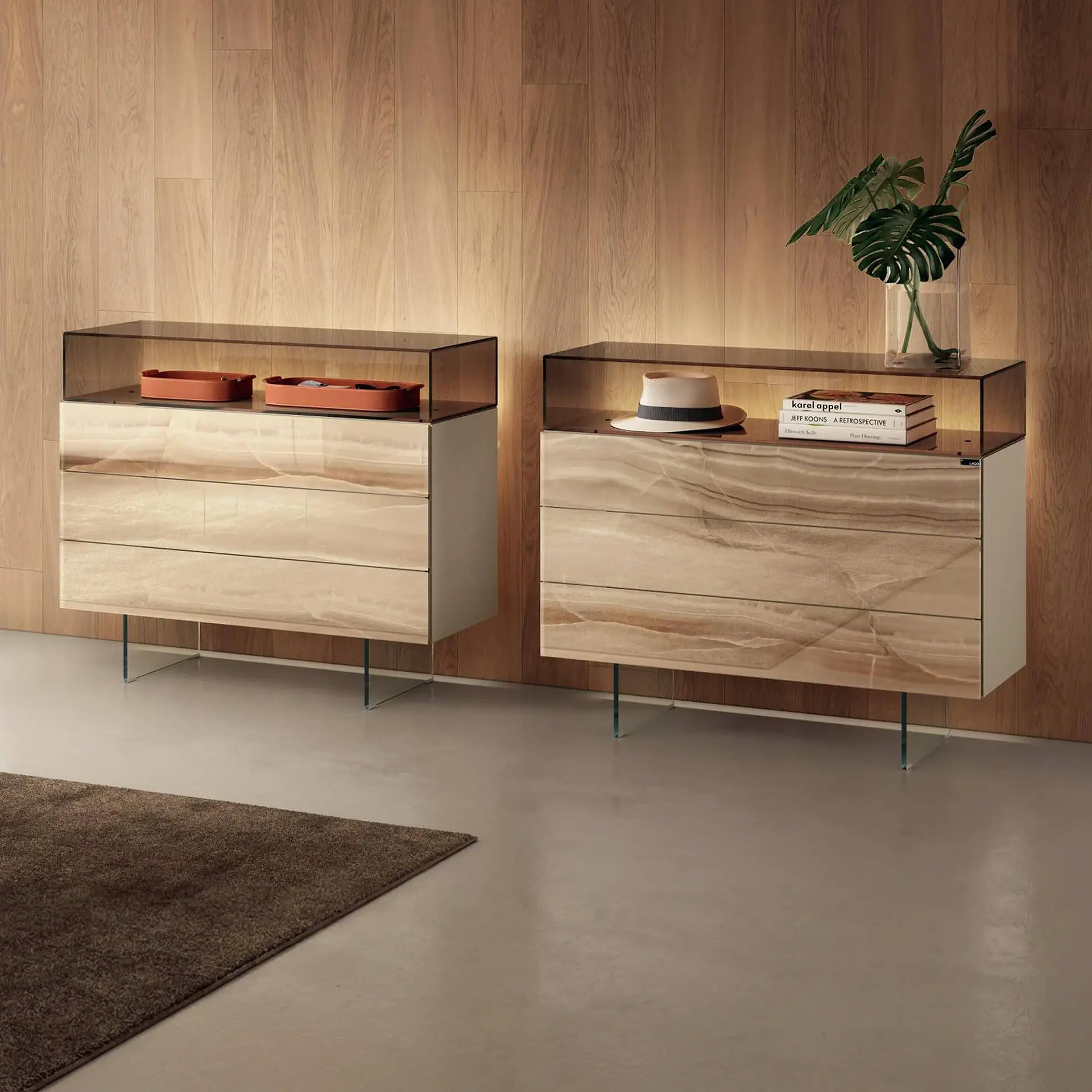 Two modern, illuminated dressers with marble-patterned drawers and clear glass legs against a light wood-paneled wall. One dresser holds decorative trays, the other a hat, books, and a vase with Monstera leaves.
