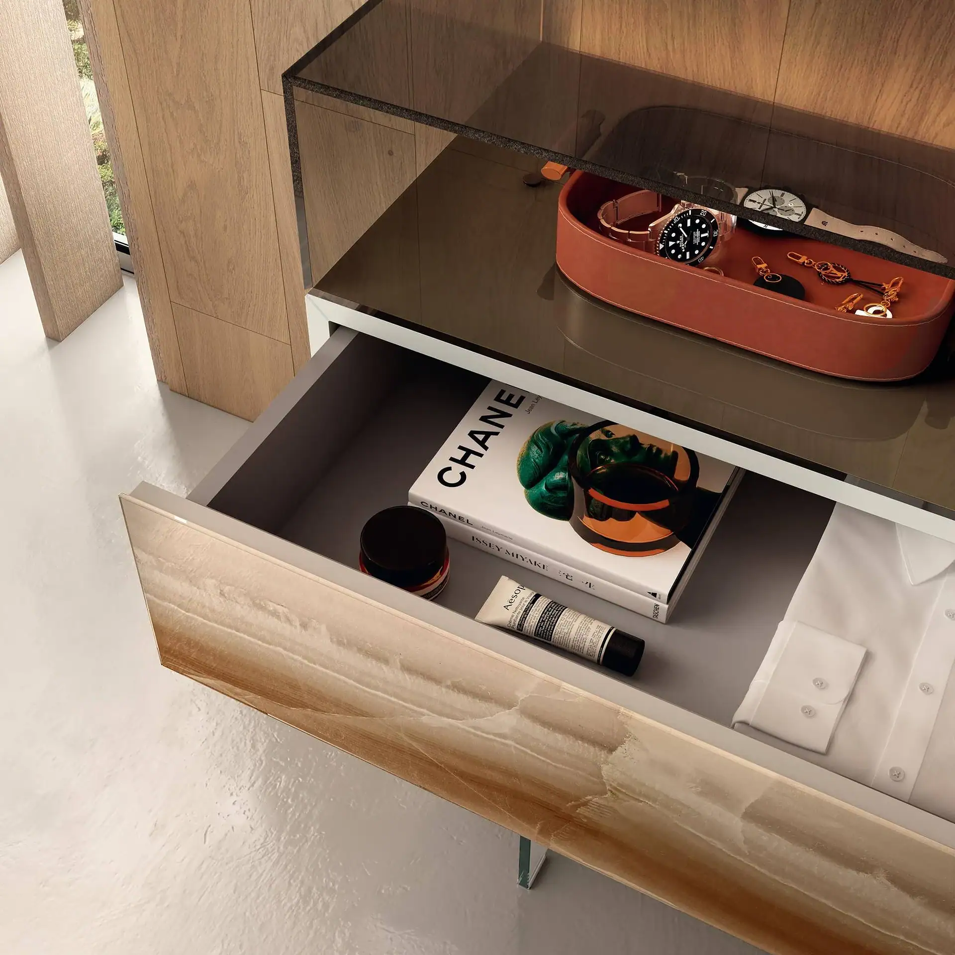 An open drawer unit with a top shelf holding watches and keys in a tray, and the open drawer containing a Chanel book, Aesop cream, and a white shirt.