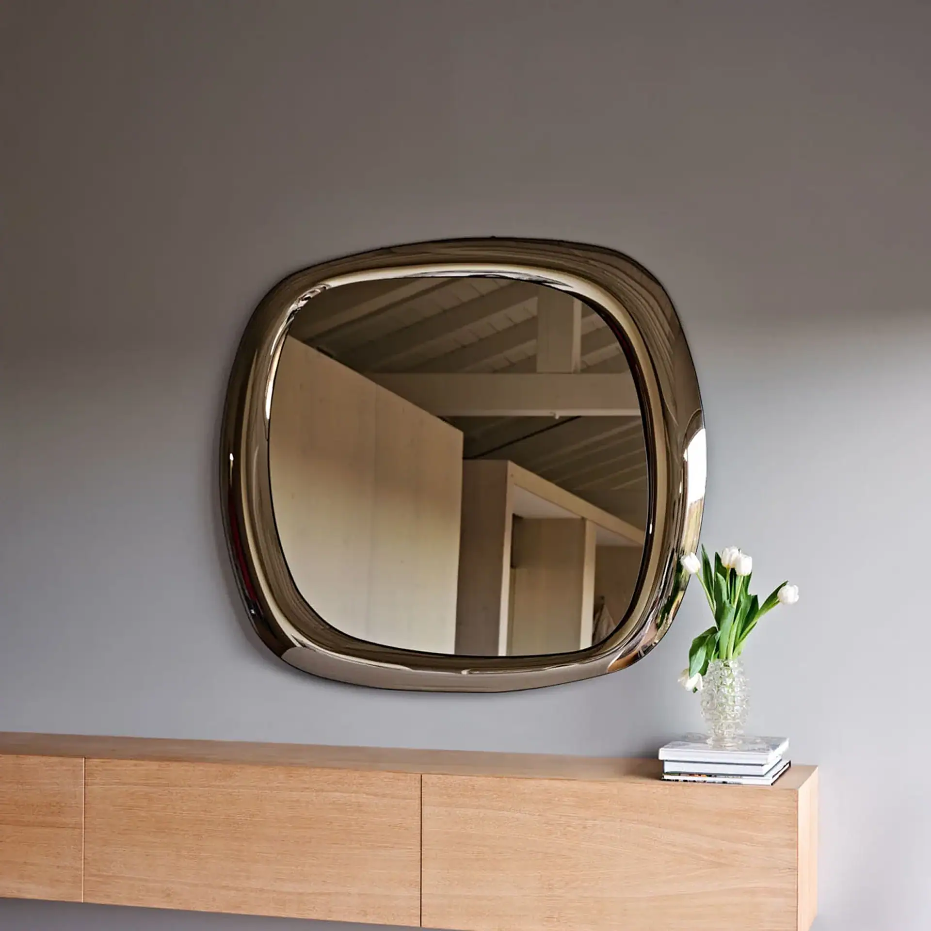 A large, irregularly shaped mirror with a glossy dark frame hangs above a light wooden floating console table. On the table, a vase of white tulips sits next to a stack of books. The mirror reflects a modern interior with wooden ceilings and beams.